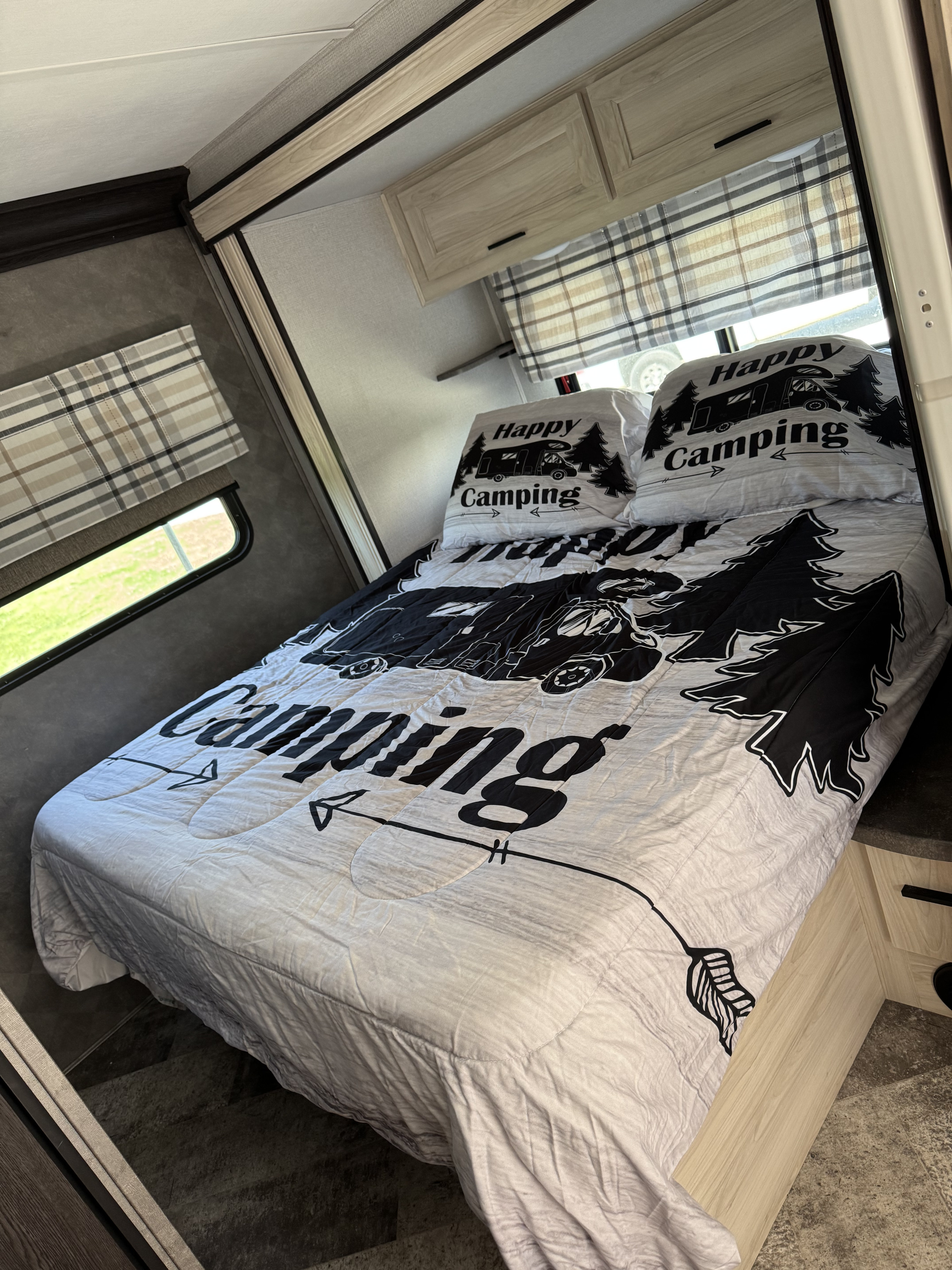 RV rental bedroom with a fun "Happy Camping" bedspread, pillows, plaid blinds, and light wood cabinetry.. Forester LE 2020