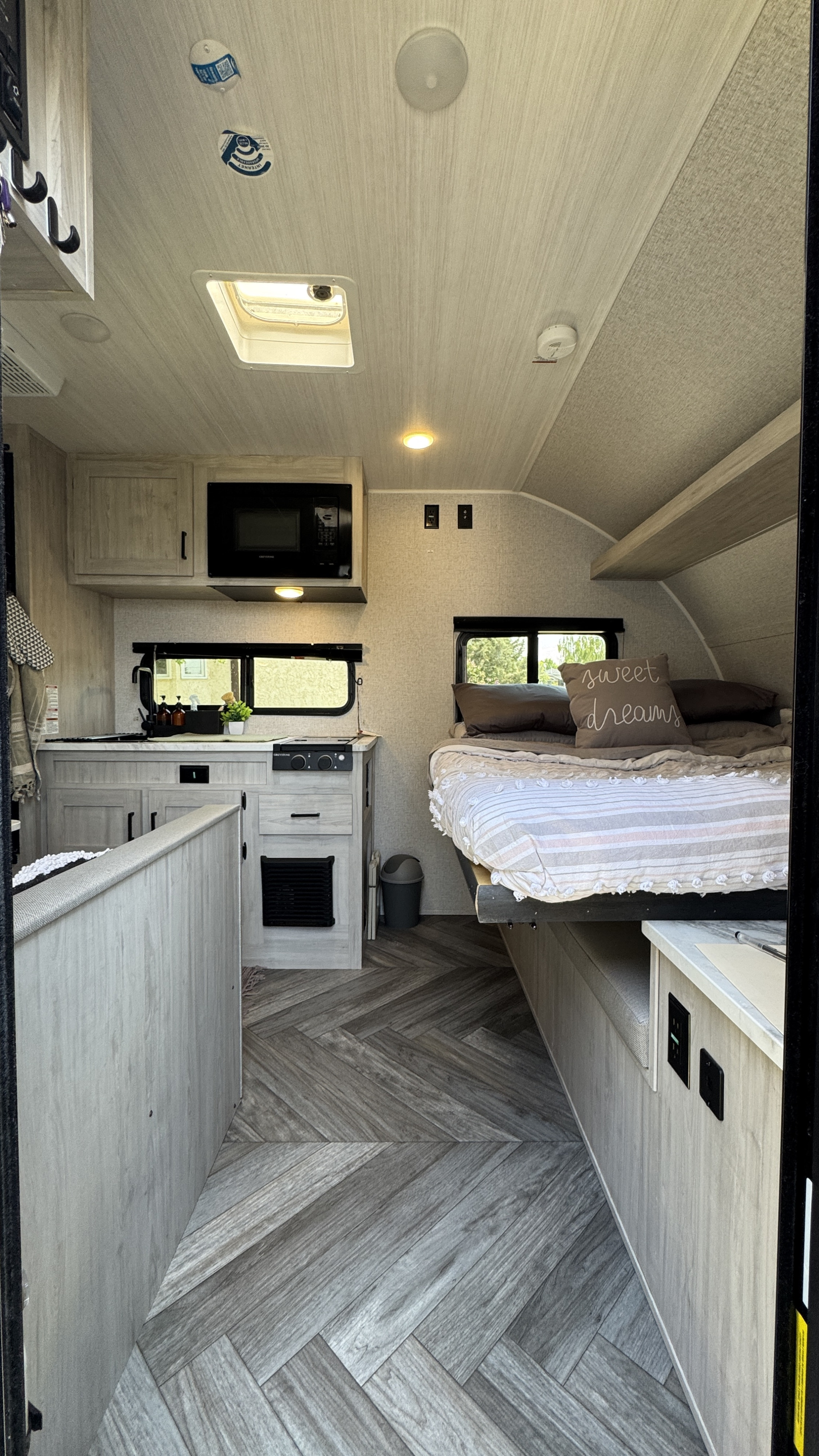 Cozy RV rental interior featuring a compact kitchen with microwave, light wood flooring, and a comfortable bed with a 'sweet dreams' pillow.. East to West Della Terra LE 2023
