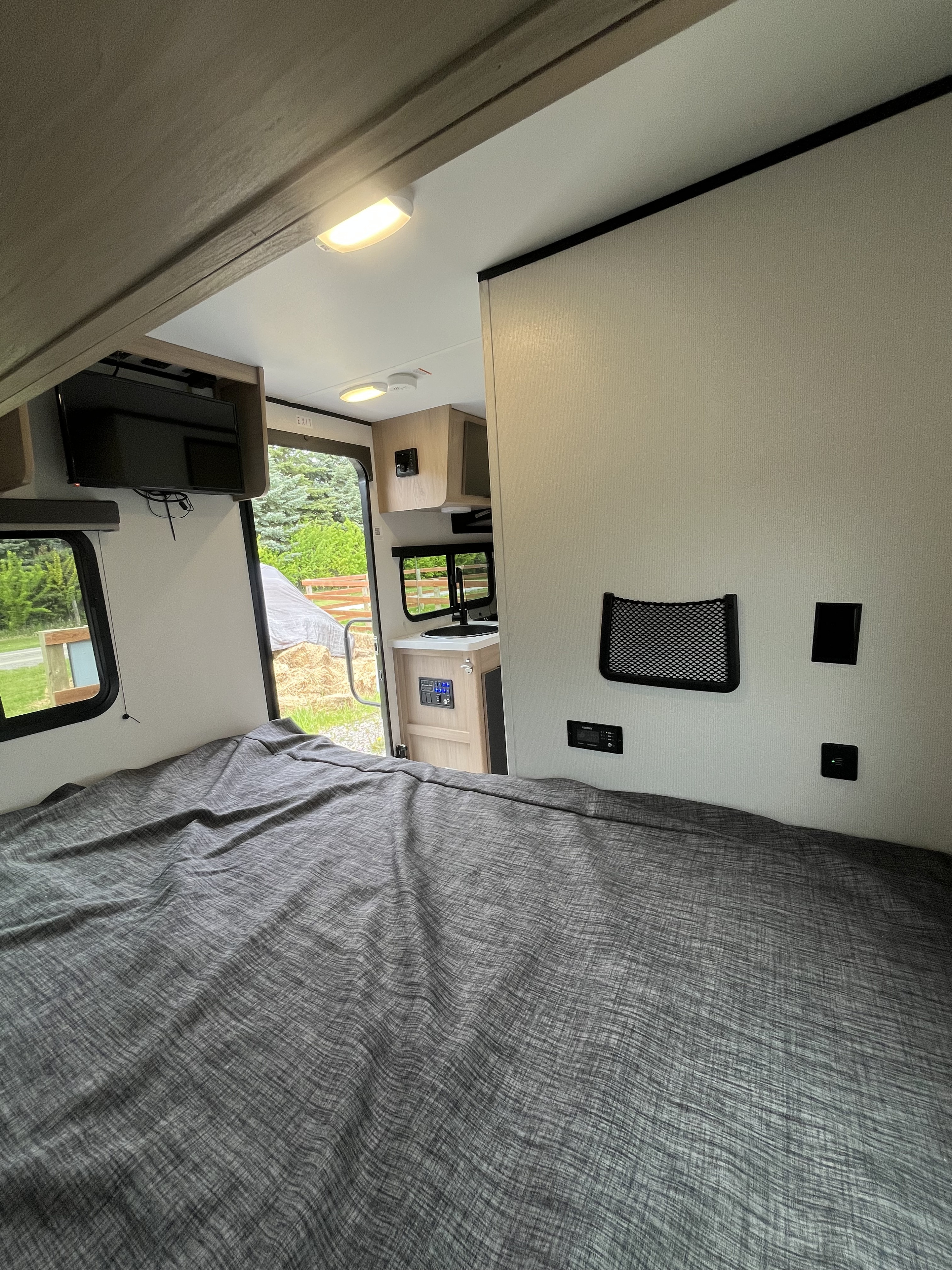 Cozy RV rental interior: bedroom view with bed, controls, TV, and kitchen area visible, looking out to nature.. Cruiser Hitch 16RD 2021