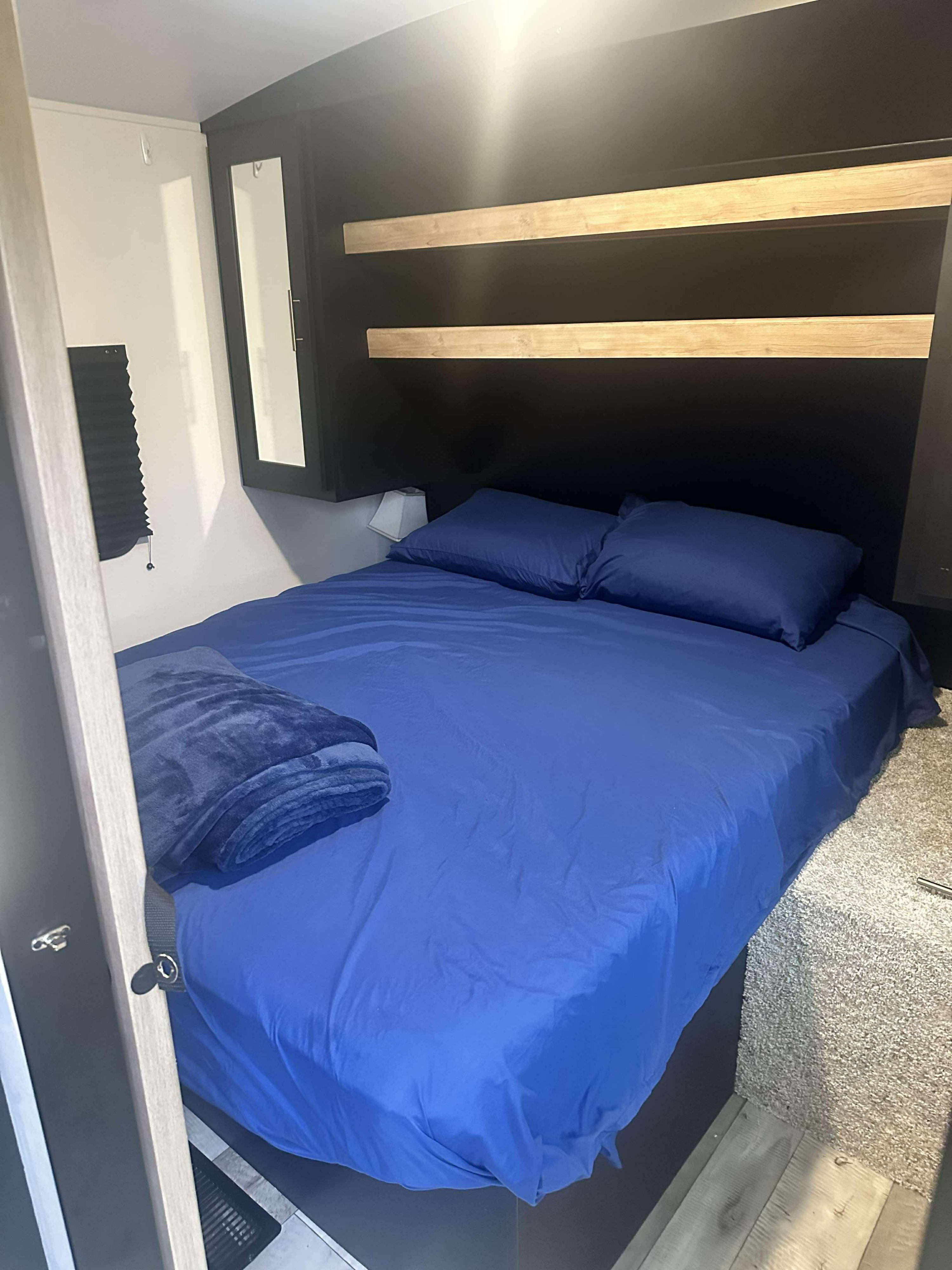 RV rental bedroom featuring a bed with blue sheets, pillows, and blanket. Dark headboard, wooden shelves, and a wall lamp visible.. Keystone RV Springdale 2022