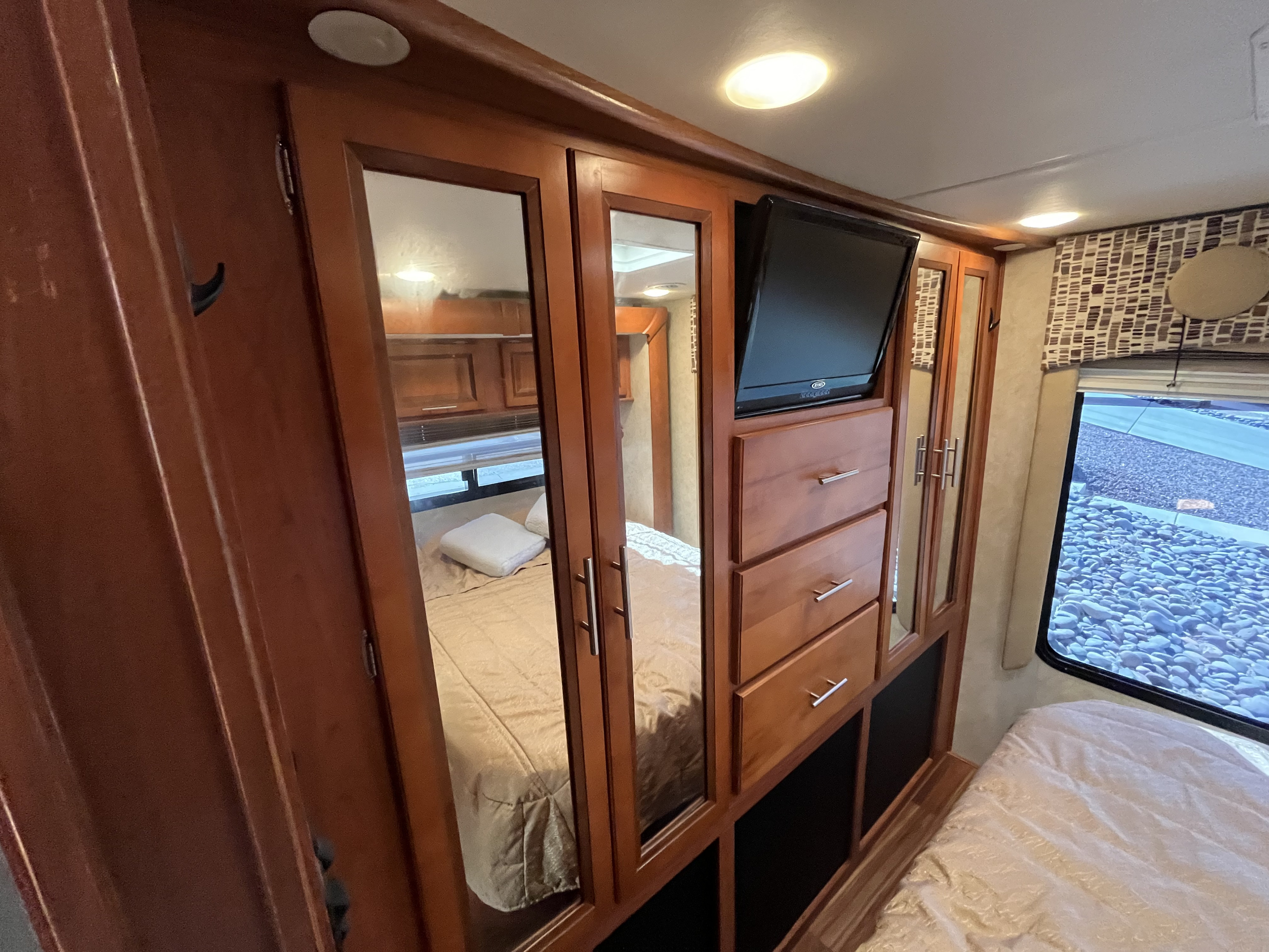 Spacious RV rental bedroom featuring a mirrored wardrobe, built-in TV, drawers, and a comfortable bed. A window shows an outdoor gravel area.. Forest River Coachmen Concord 2013