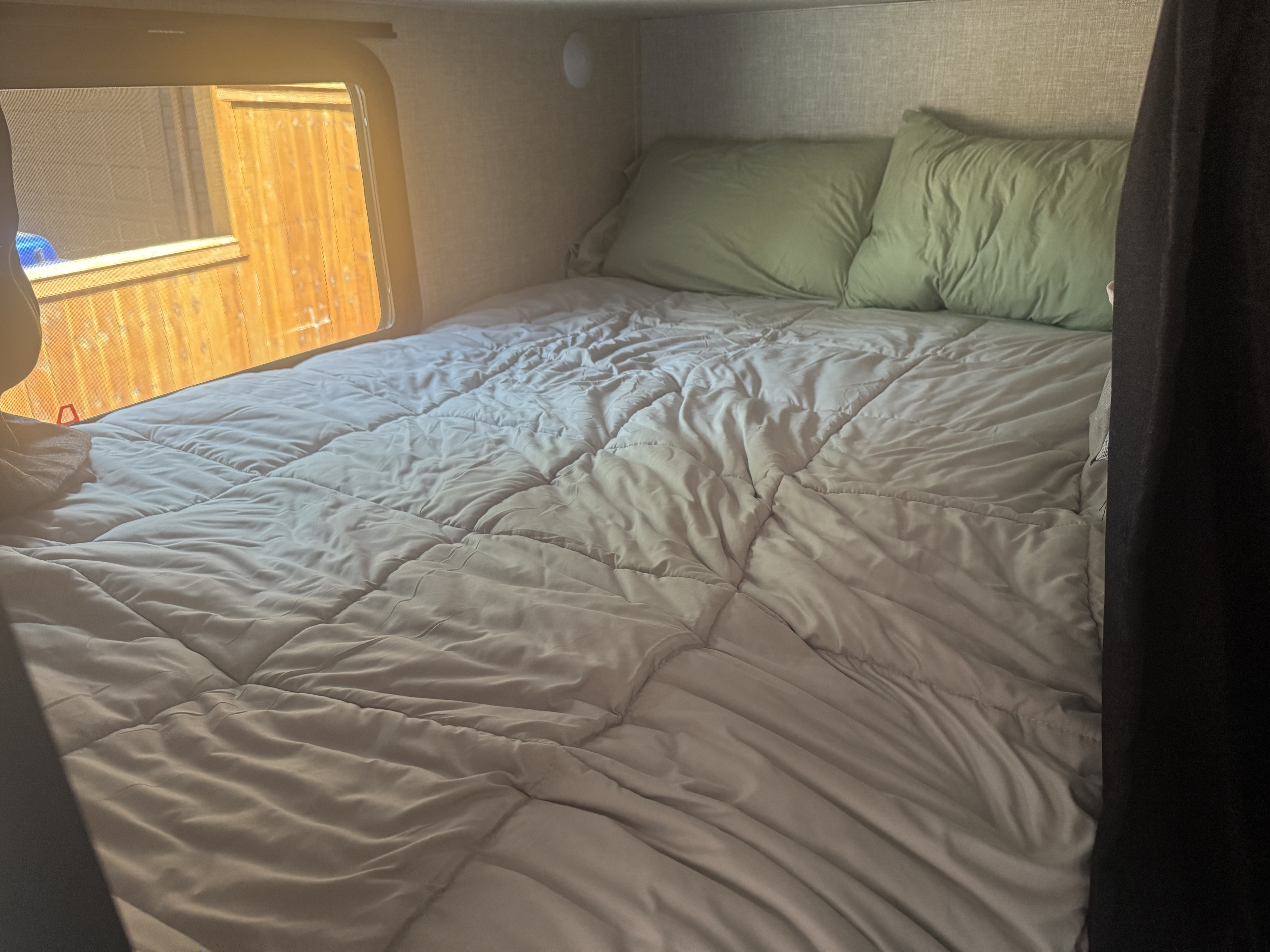 Comfortable RV rental bed with light grey comforter & green pillows, next to a window showing a wooden fence.. Jayco Jay Flight SLX Rocky Mountain Edition 2022