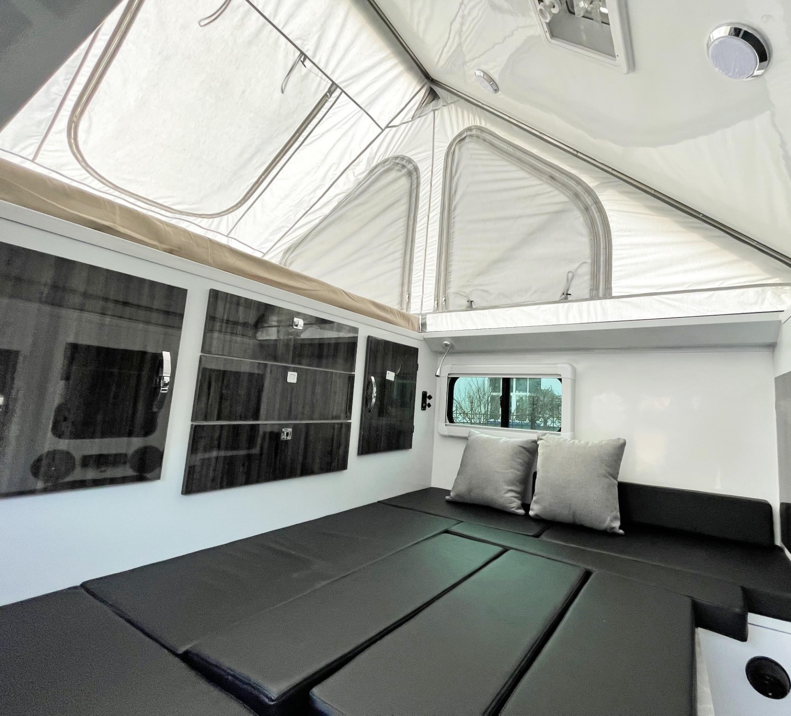 Inside a modern RV rental: a bright pop-up roof, black padded seating that converts to a bed, and wall storage.. Zoomies Campers Spirit 2025