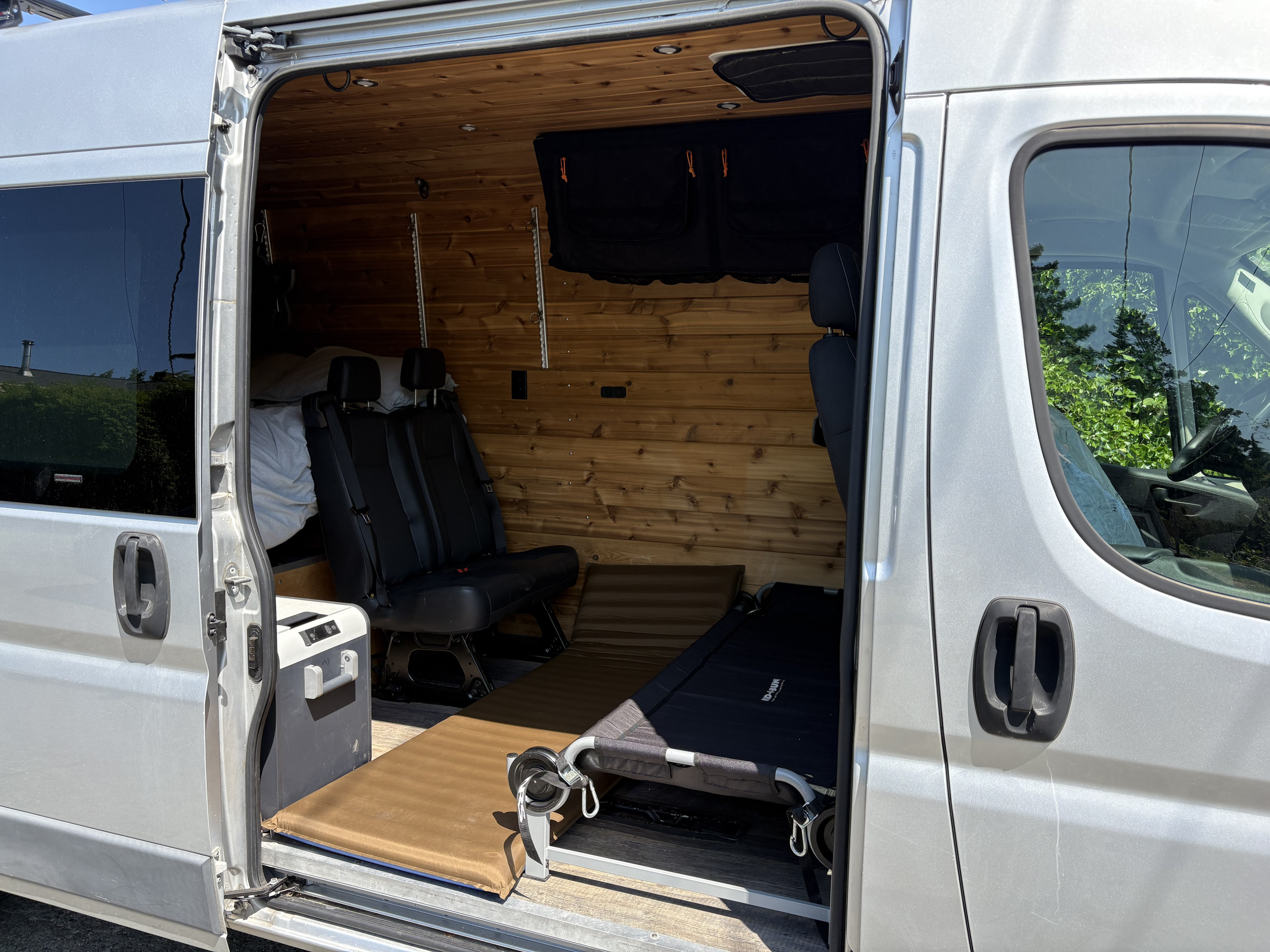 Inside this silver RV rental, wood paneling, black seats, a cooler, sleeping pad, and a cot offer adventure-ready comfort.. Ram Promaster 2500 2018