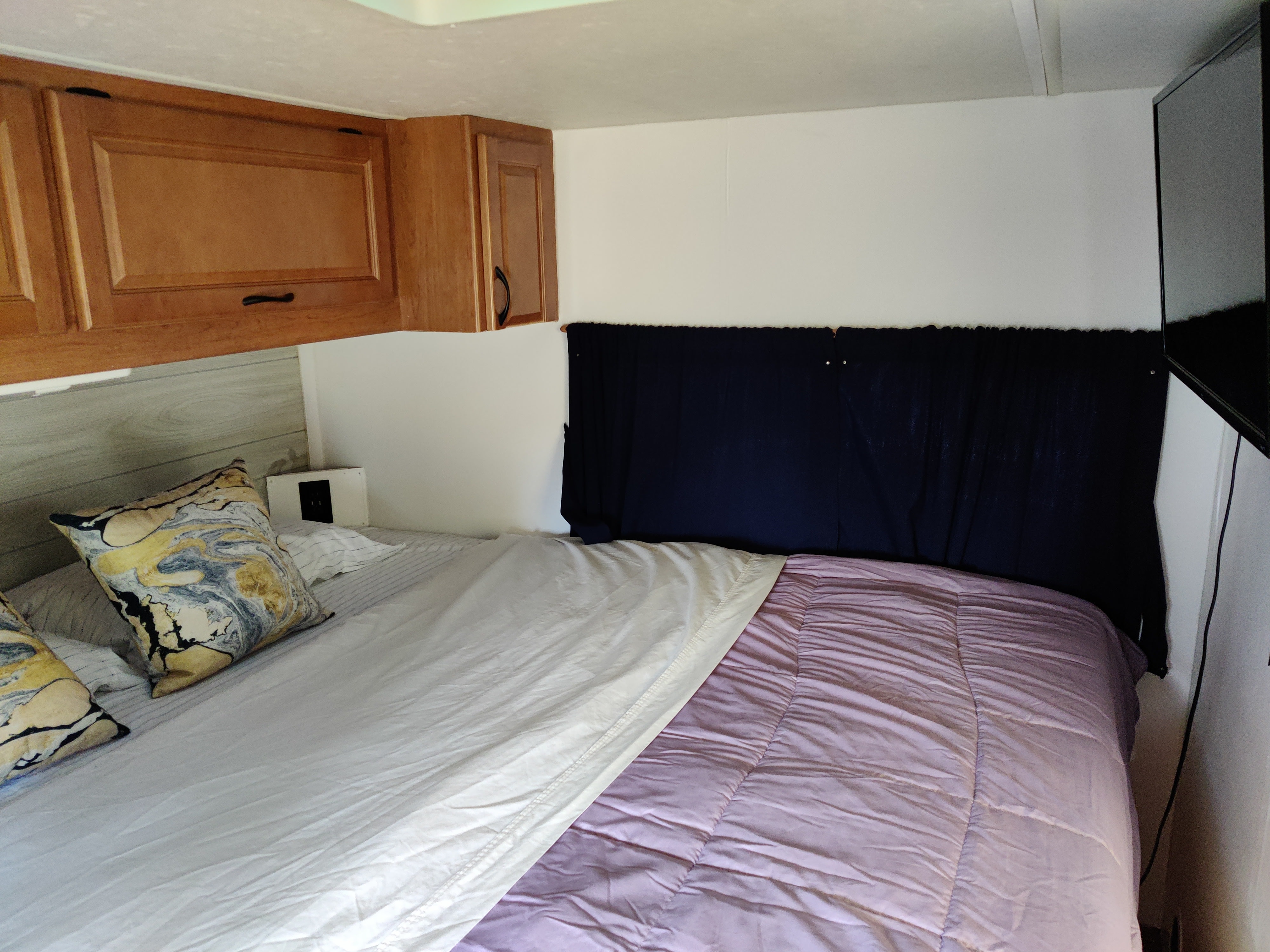 RV rental bedroom: Features include wooden overhead cabinets, a bed with a light purple comforter & patterned pillows, a fabric headboard, & TV.. Thor Majestic 2015