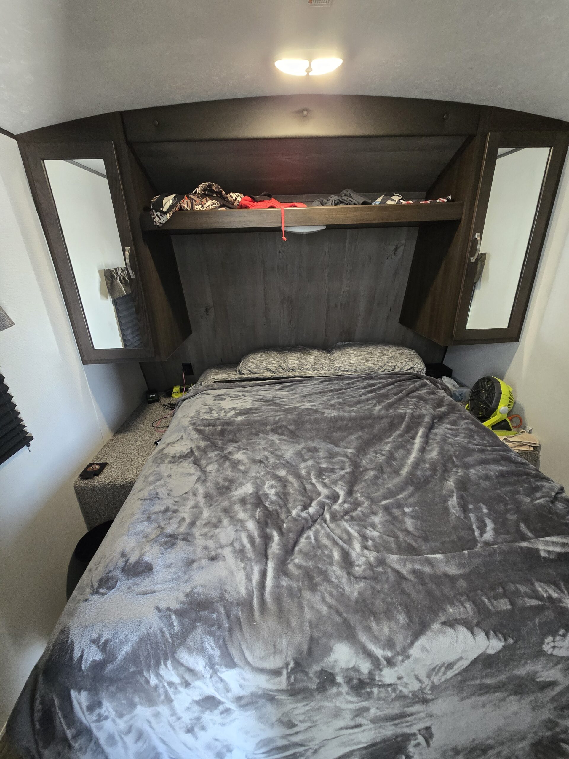 Cozy RV rental bedroom with a full bed, gray bedding, overhead storage cabinets, and a bright ceiling light. Ideal for camper trips.. Keystone Springdale 2024
