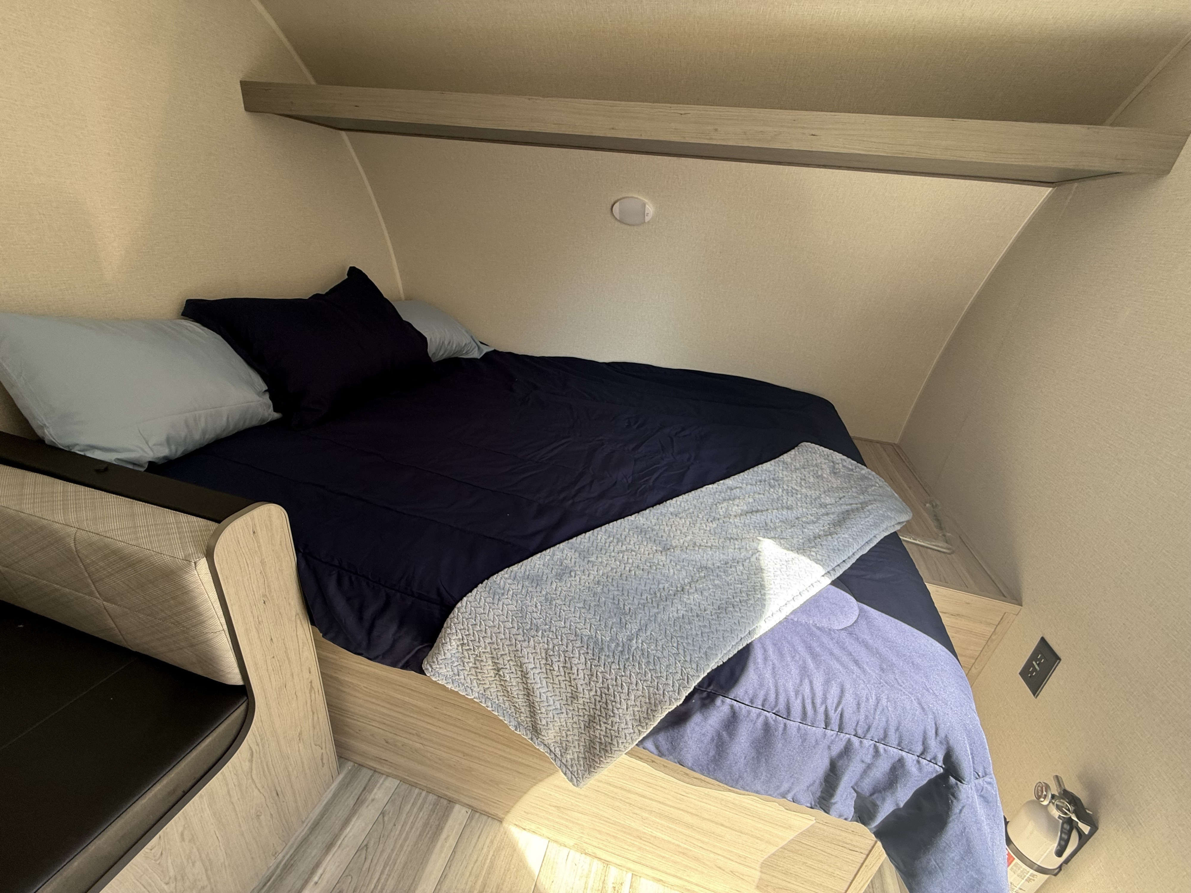 RV rental bedroom with a cozy bed, blue bedding, overhead shelf, bench, and fire extinguisher for safety.. Heartland Pioneer 2025