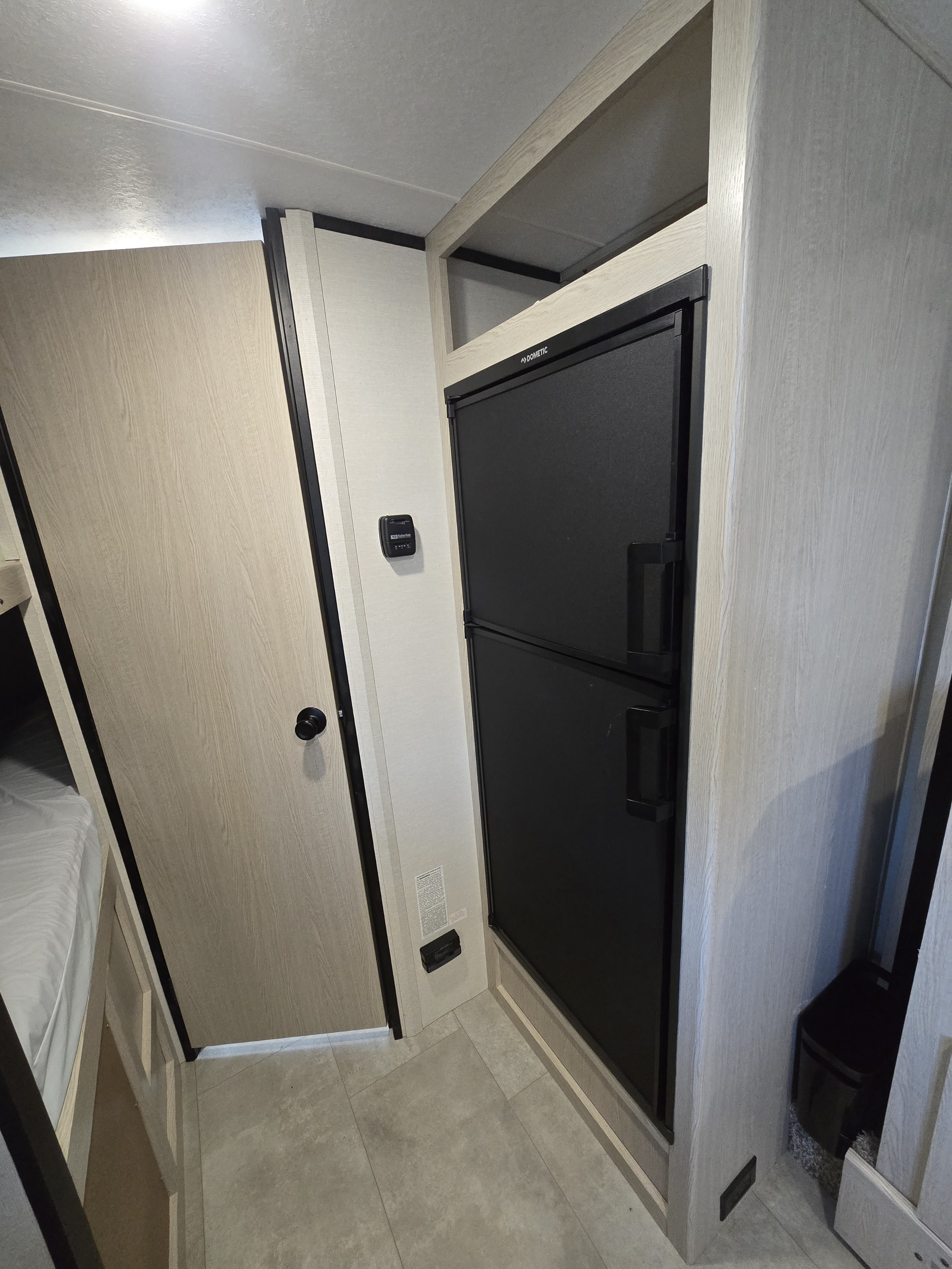 Interior of an rv rental, featuring a black Dometic fridge, a light wood door, and a glimpse of a bunk bed.. Coachmen Apex Nano 2021