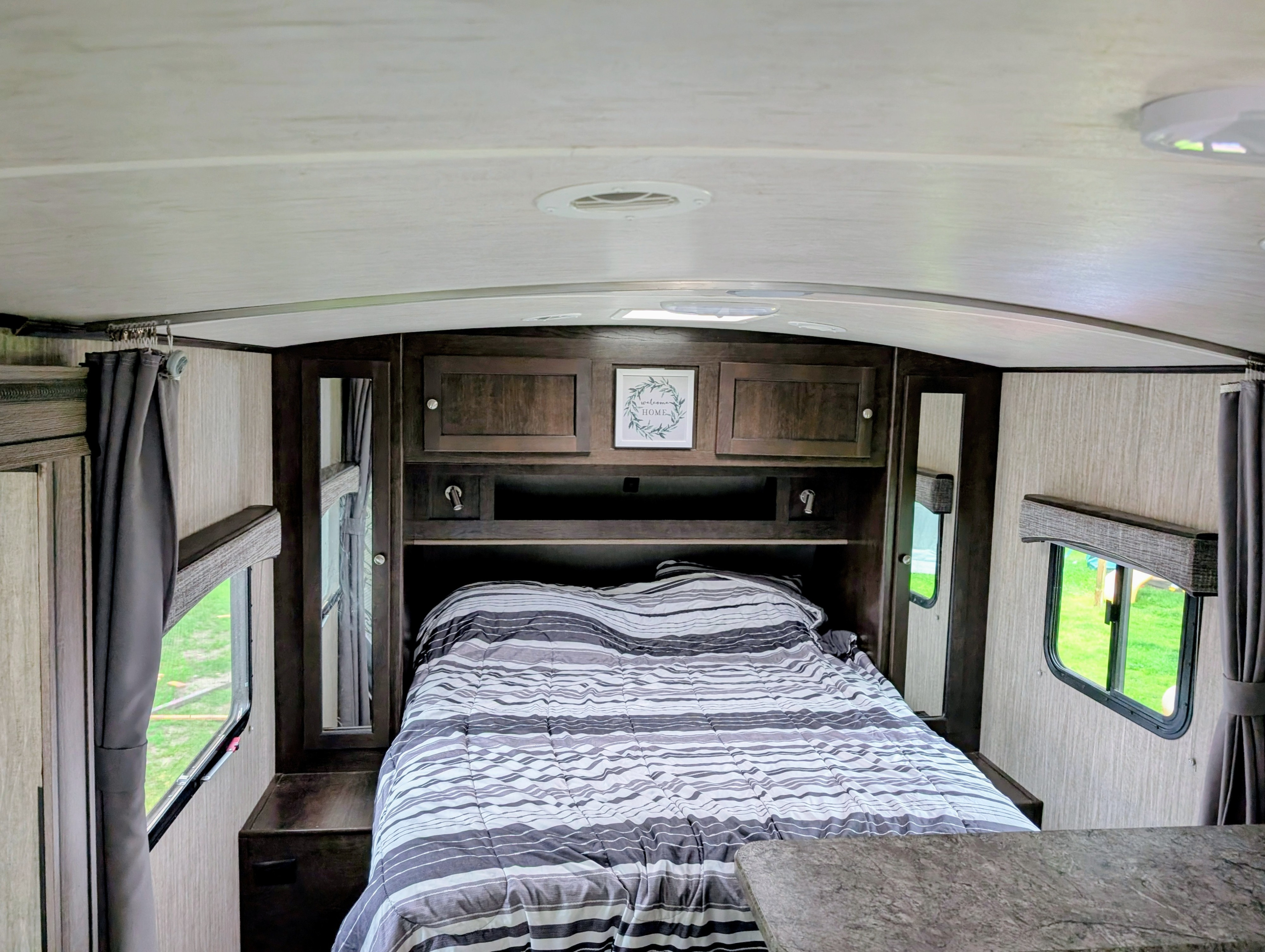 This RV rental bedroom features a compact, cozy interior with a striped bed, overhead cabinets, and windows showing green outdoors.. Heartland RVs Sundance 2019
