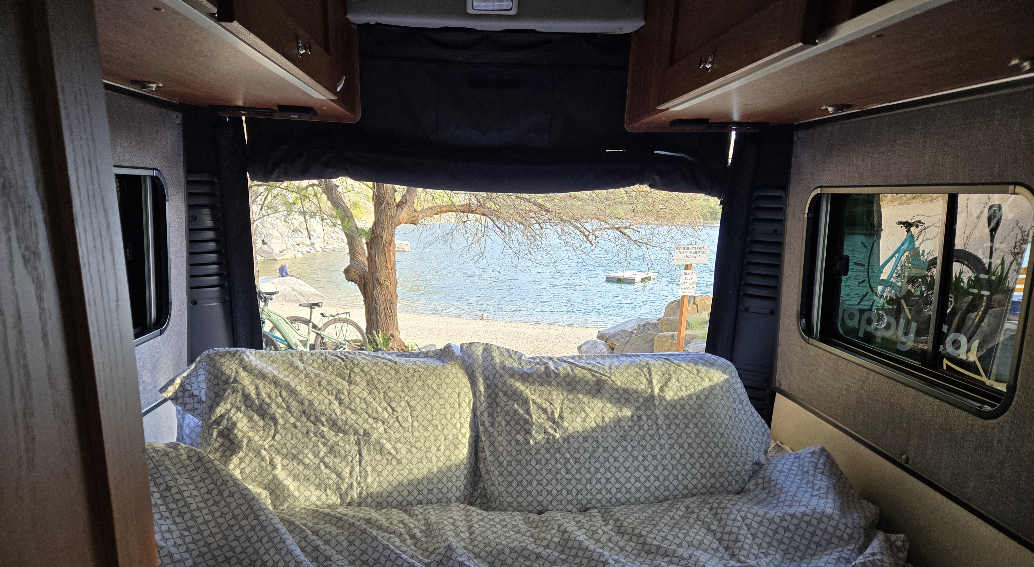 Relaxing in an RV rental, looking out at a stunning lake view from the cozy rear bed. Ideal for peaceful camping.. Roadtrek Roadtrek Class B 2021
