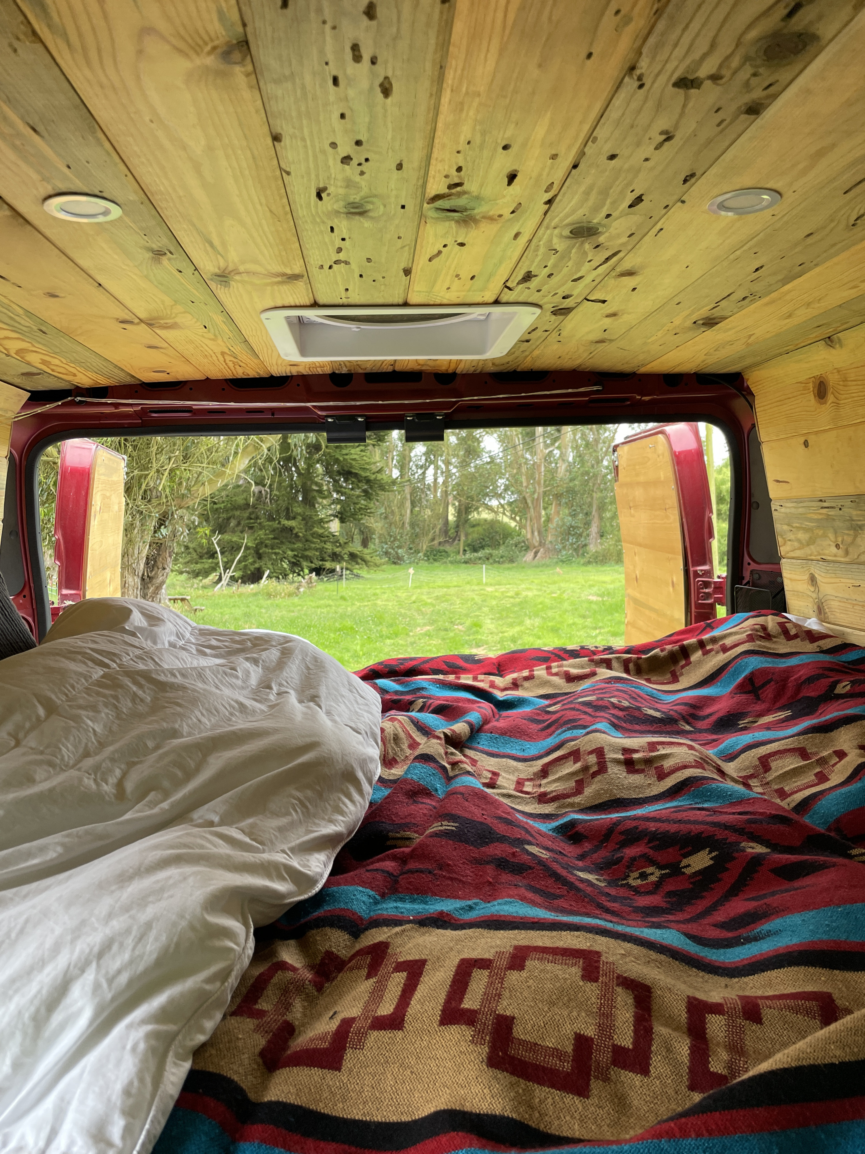 Inside a cozy RV rental: a bed with patterned blanket, wooden ceiling, and open rear doors revealing a serene green field.. Dodge Ram Promaster 2020