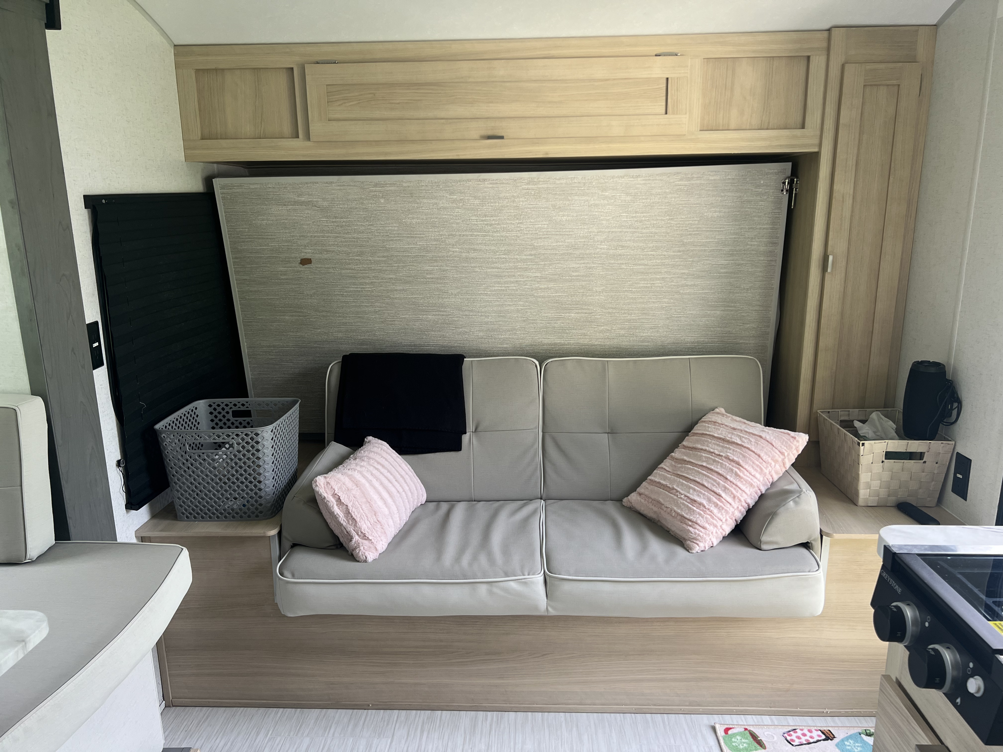 Cozy RV rental living area featuring a light gray sofa, hidden murphy bed, and light wood overhead cabinets. Storage baskets also seen.. Forest River Salem FSX 2023