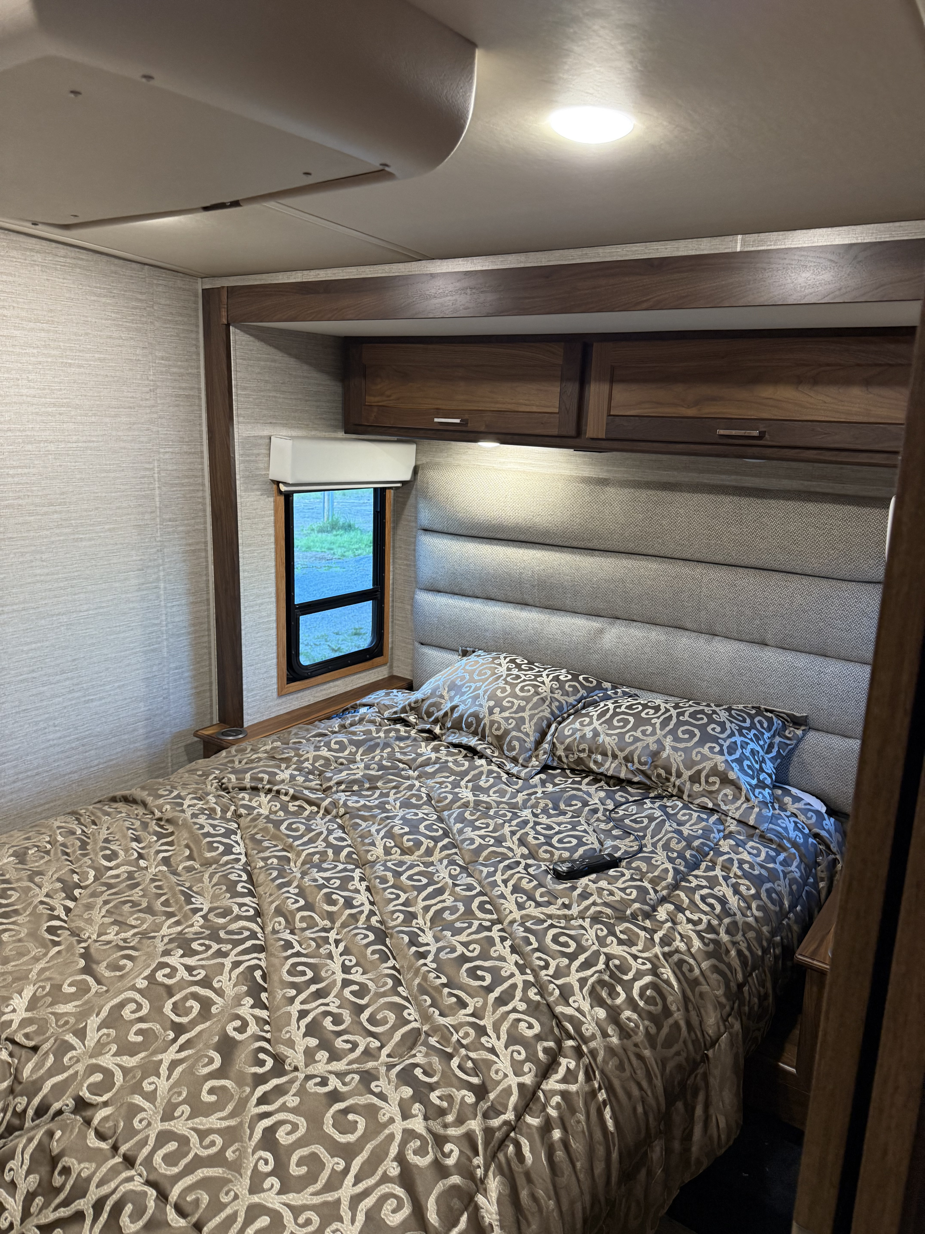 Cozy RV rental bedroom featuring a patterned bedspread, padded headboard, window, and overhead cabinets for a comfortable stay.. Winnebago Forza 2021