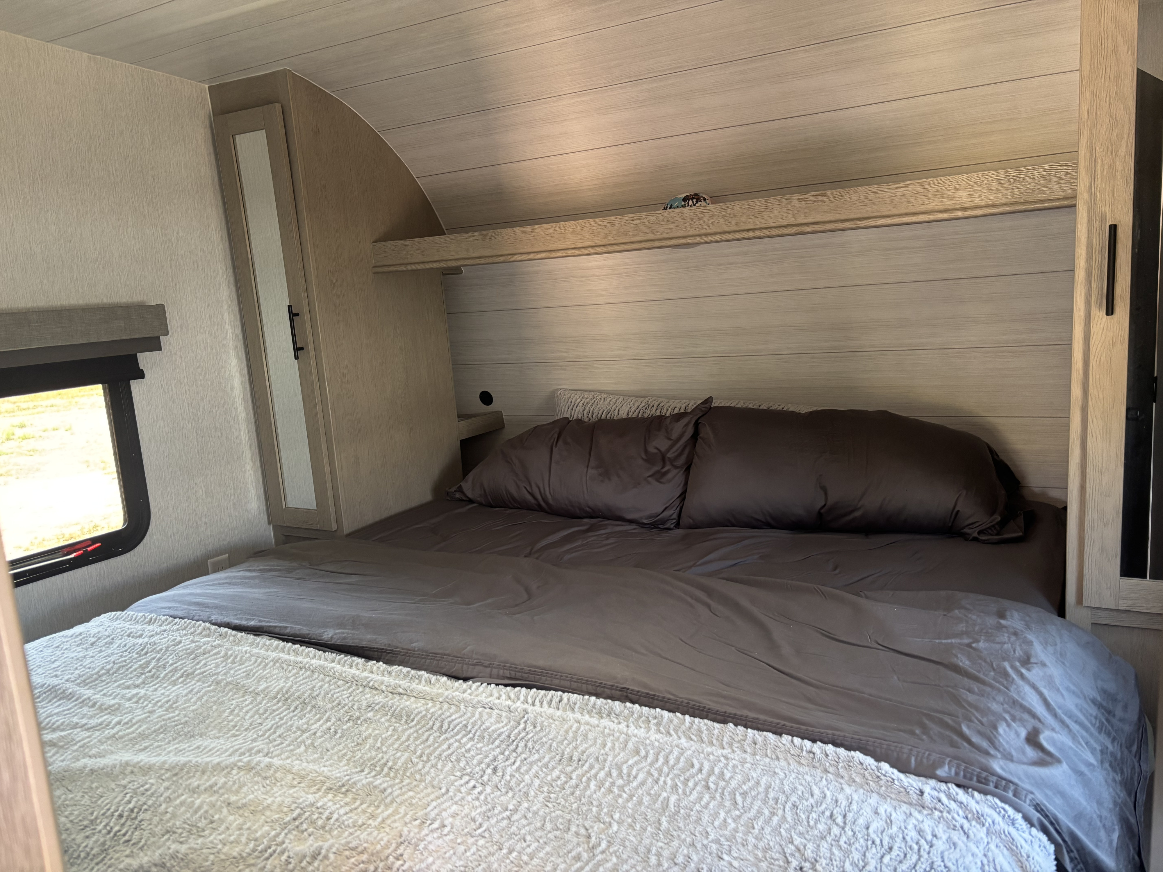 Cozy RV rental bedroom with dark bedding, white blanket, and wood accents. Perfect compact interior for travel.. Forest River Stealth 2024