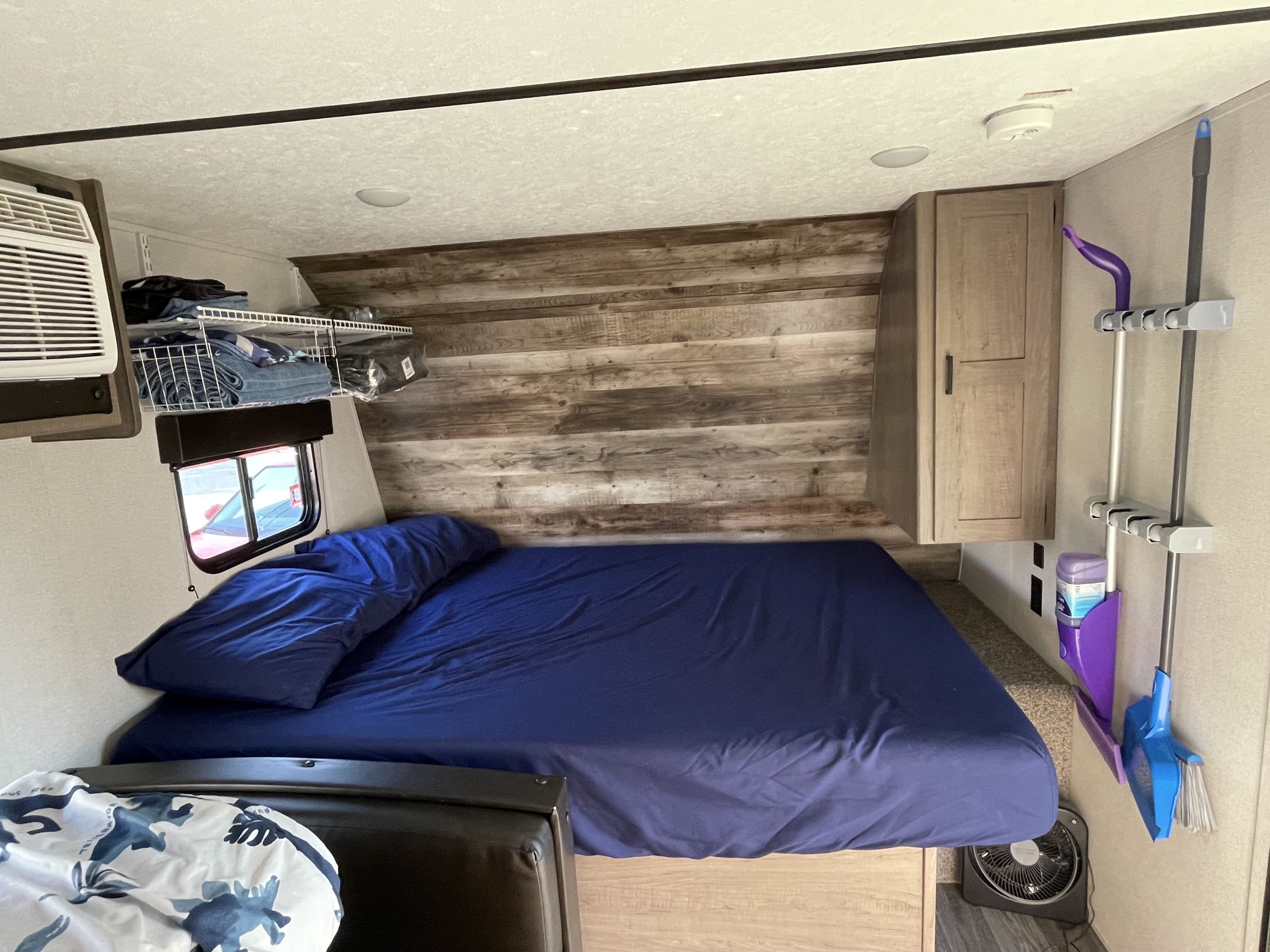 This RV rental bedroom features a comfy blue bed, rustic wood accent wall, practical storage, AC unit, and cleaning supplies.. Keystone RV Hideout 2021