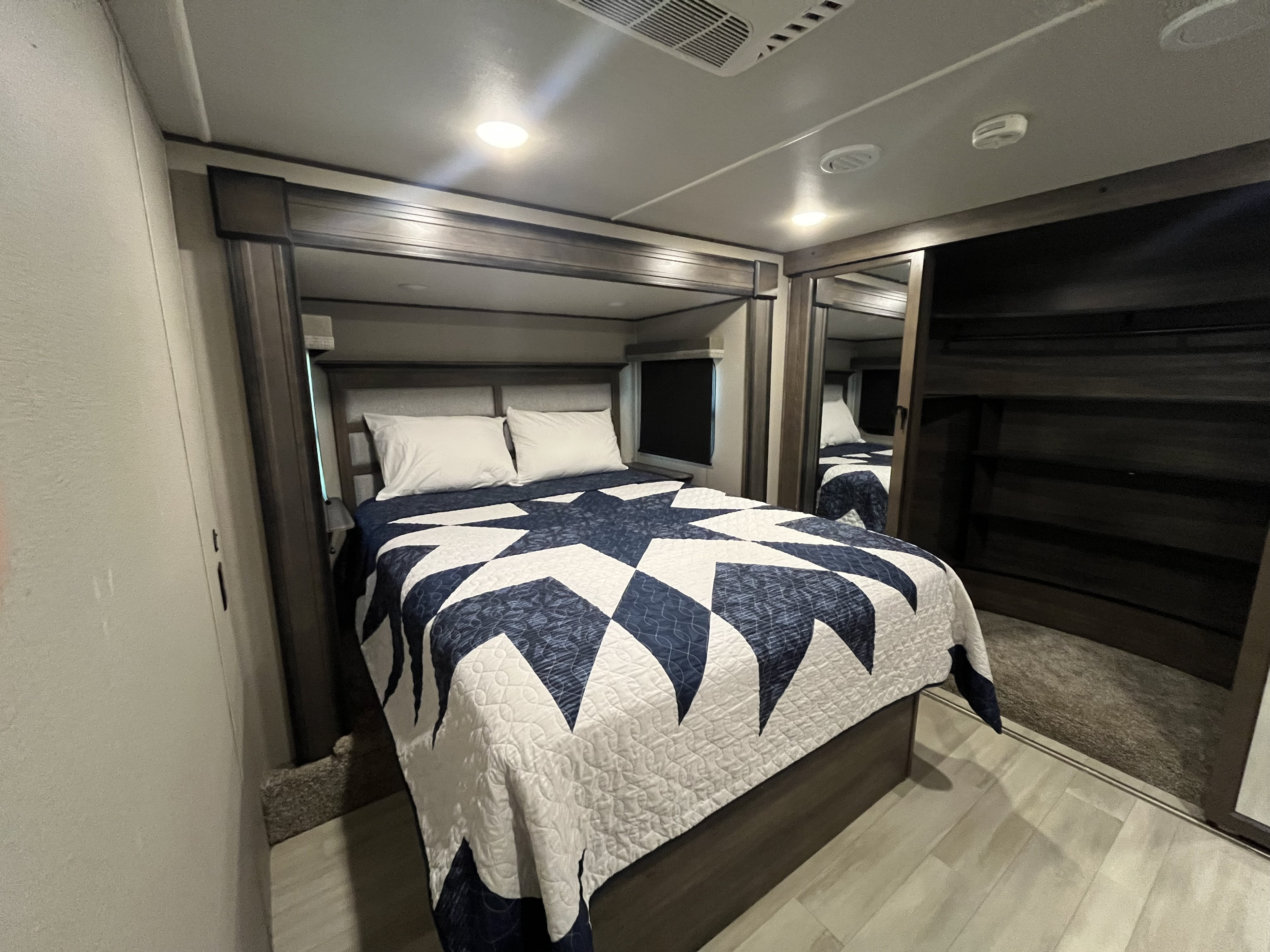 This RV rental's bedroom offers a cozy bed with a striking blue & white quilt, overhead lighting, and mirrored wardrobe storage.. Grand Design Solitude S-Class 2021