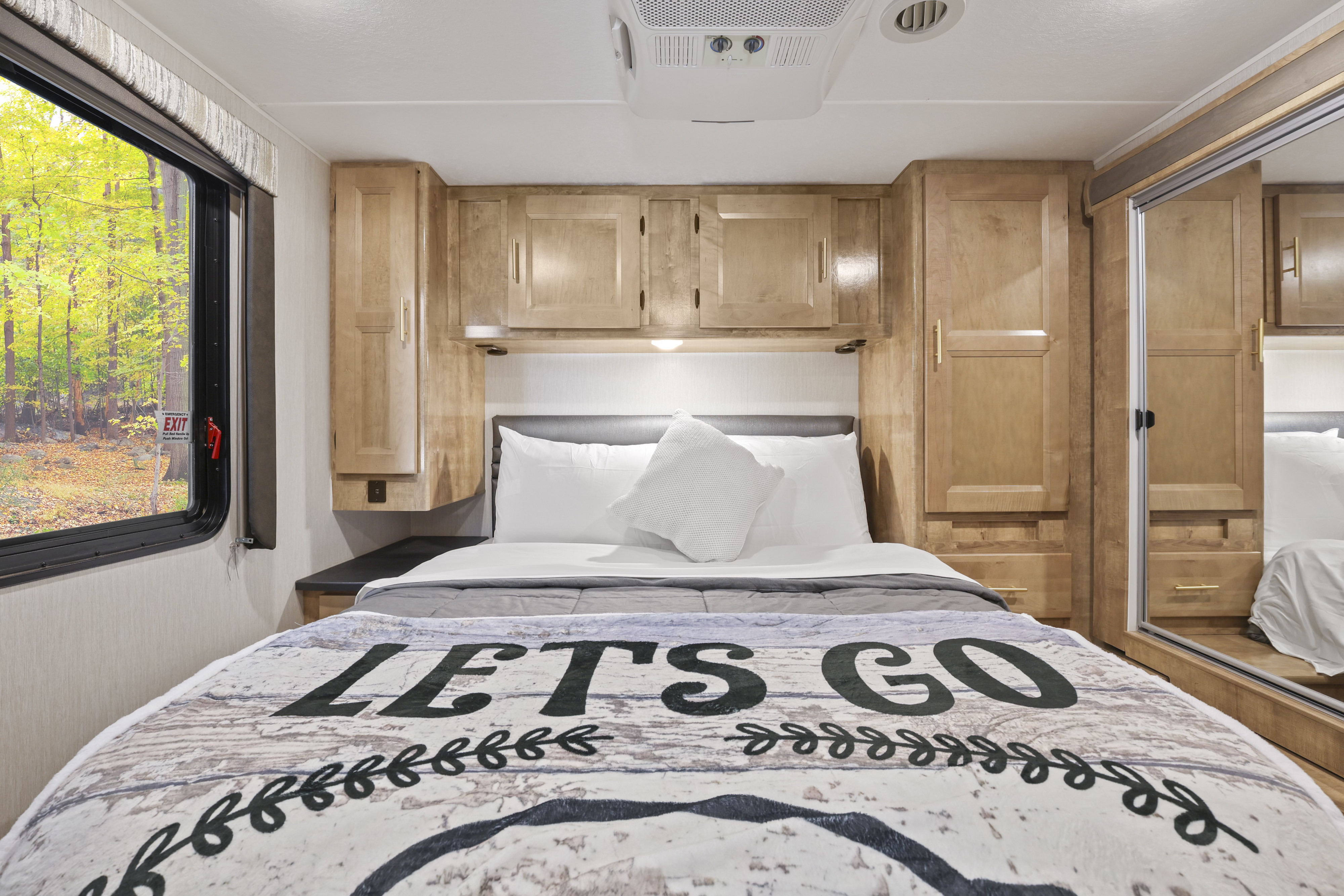 Cozy RV rental bedroom with a "LET'S GO" blanket, wood cabinets, and a scenic forest view from the window.. Forest River Coachmen Leprechaun Premier 2021