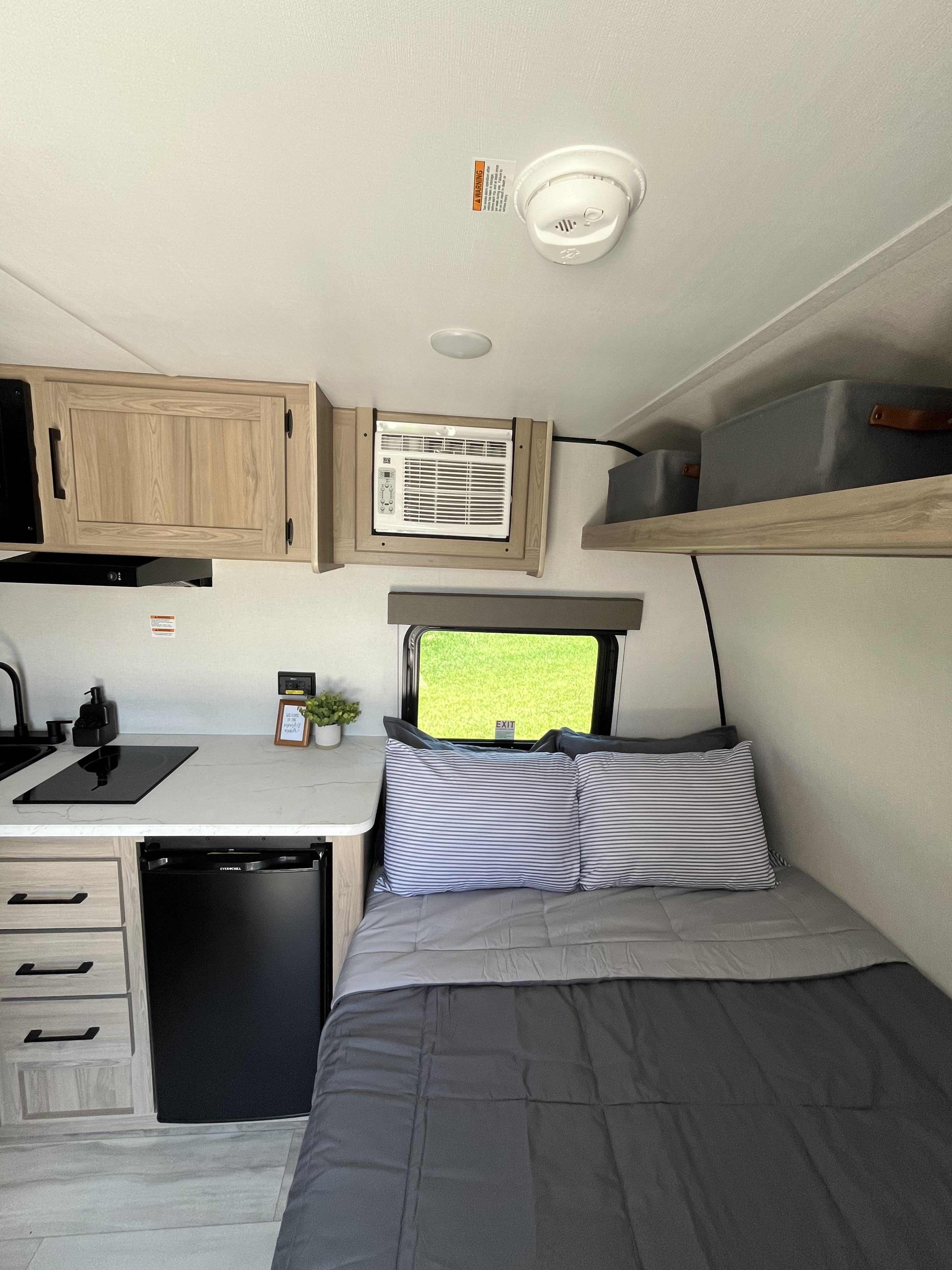 Compact RV rental interior showcasing a kitchen with sink, stovetop, and fridge, alongside a cozy bed with gray bedding. An AC unit is overhead.. Keystone Bunkhouse 2025