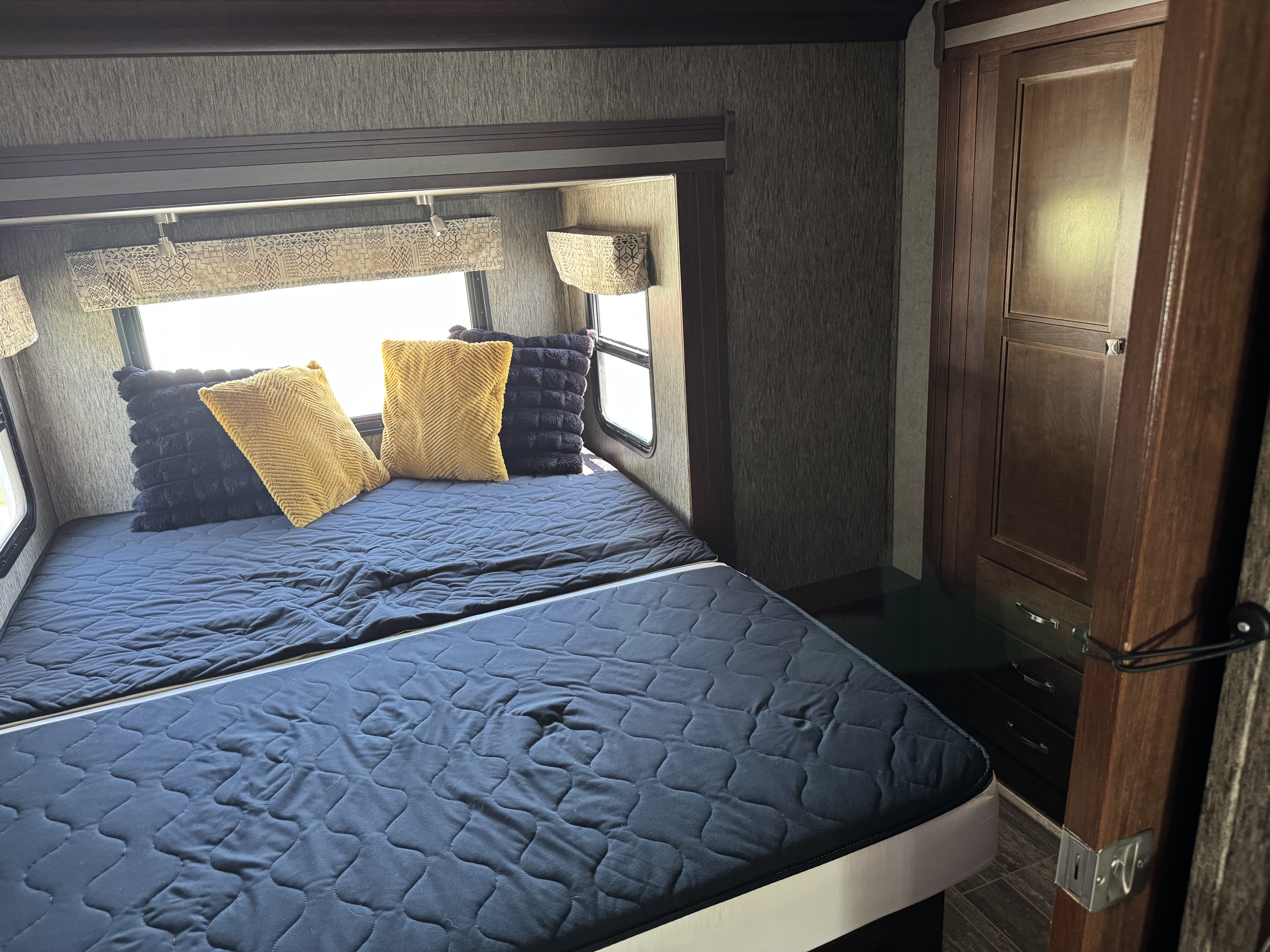 RV rental's cozy bedroom features a quilted dark blue bed, bright pillows, and wooden storage. Ready for your adventure.. Forest River Sunseeker 2018