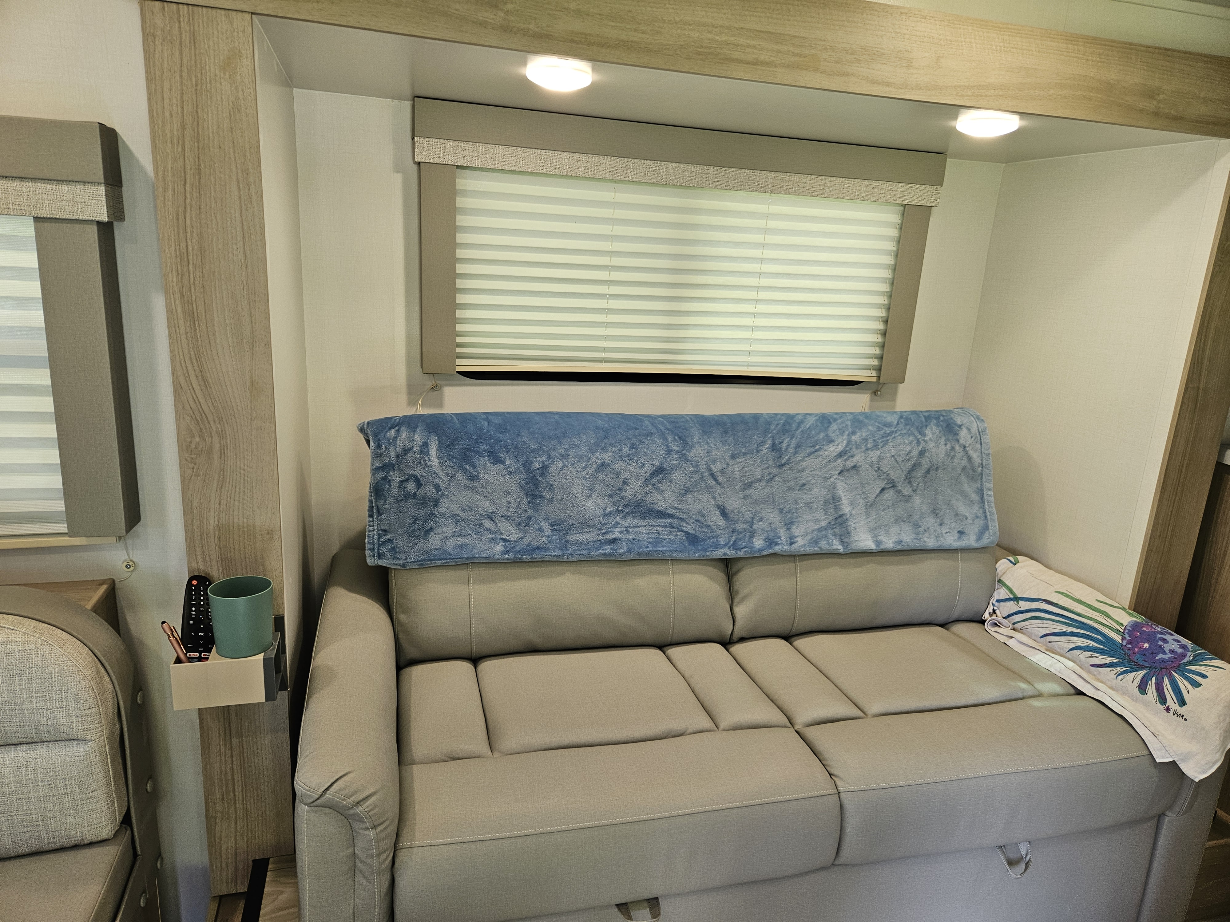 RV rental interior: light grey convertible sofa with blue blanket, window blinds, wood-look trim, and handy wall organizer.. Winnebago Minnie Winnie 2024