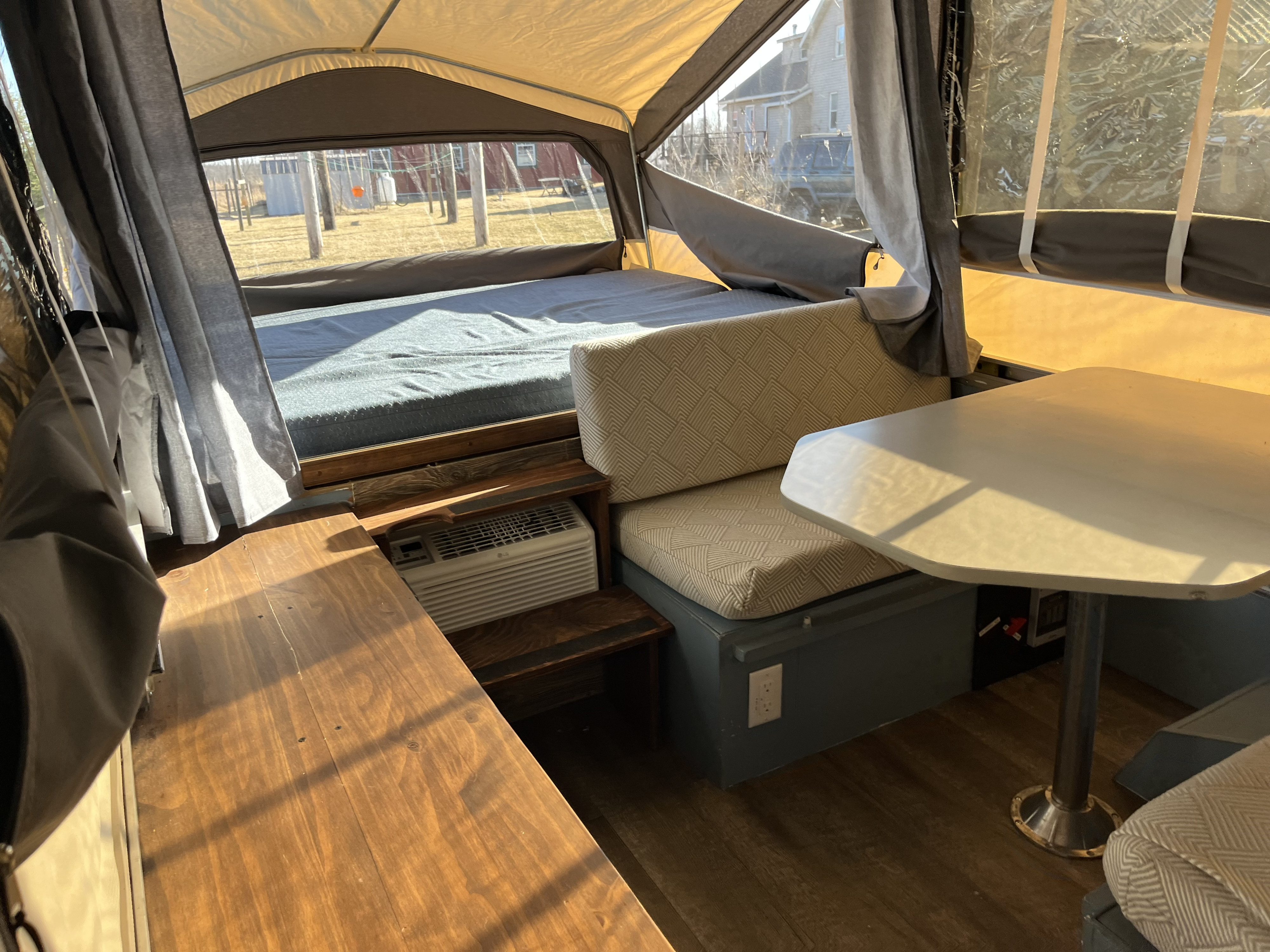 Experience this RV rental! Interior of a pop-up camper with a comfy bed, dinette, and AC unit. Wood accents & outdoor views.. Aero Coach Coach 2002