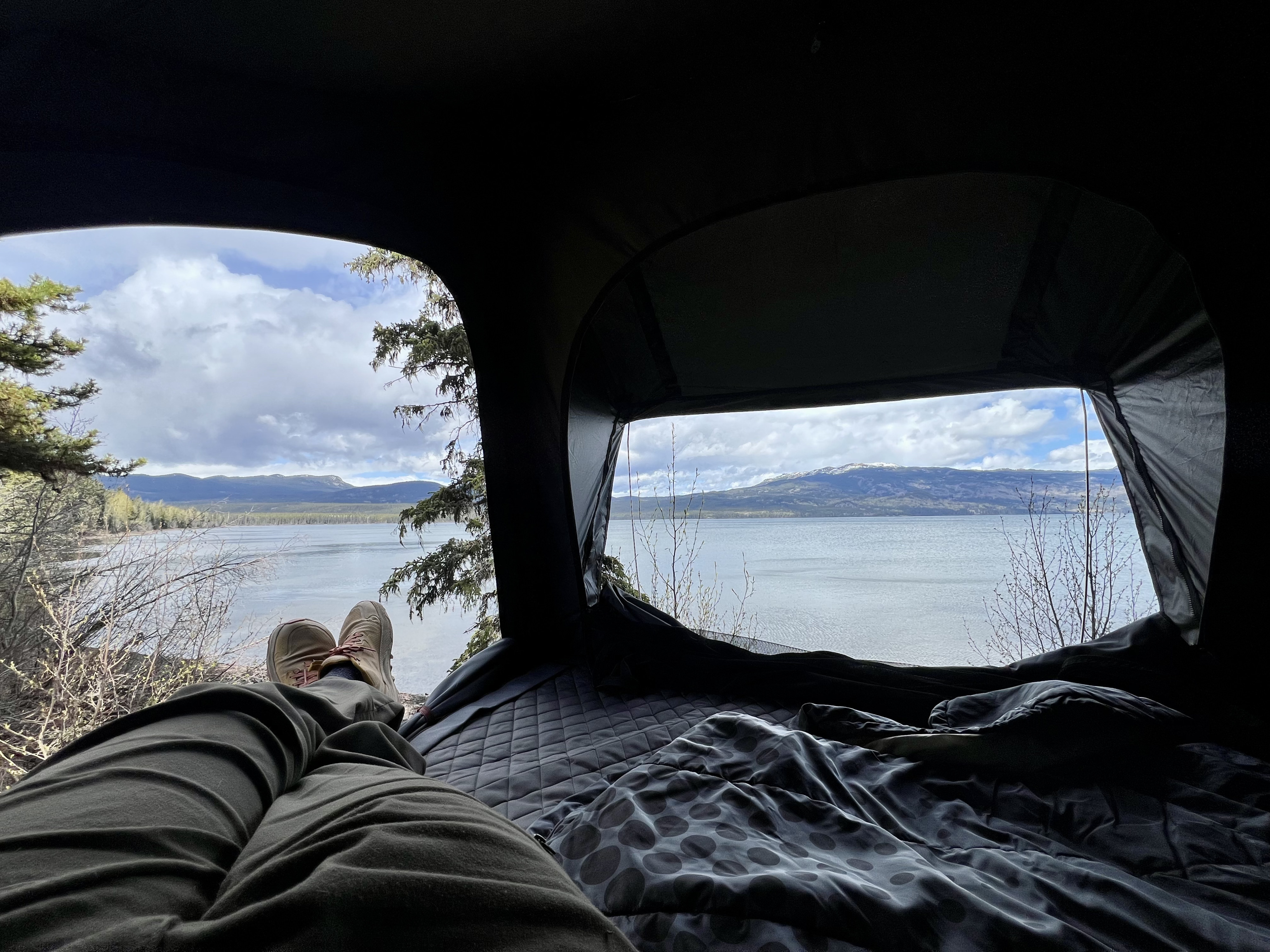 View from inside an RV rental: a person's legs stretched towards a calm lake with distant mountains and trees.. Toyota Tacoma 2020