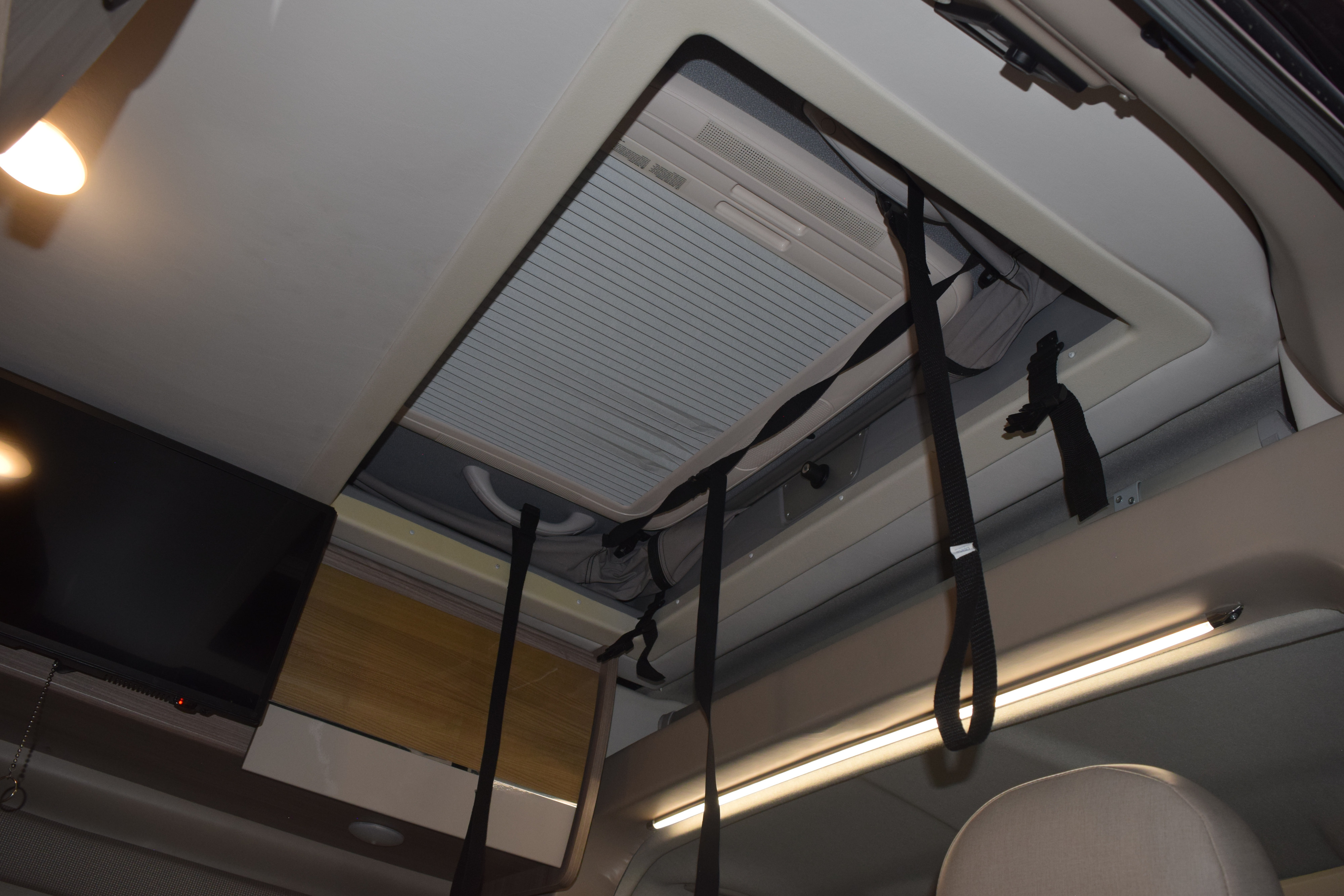 Inside an RV rental: Looking up at a large skylight with AC, mounted TV, and overhead storage cupboards with LED lighting.. Thor Sequnce 2023