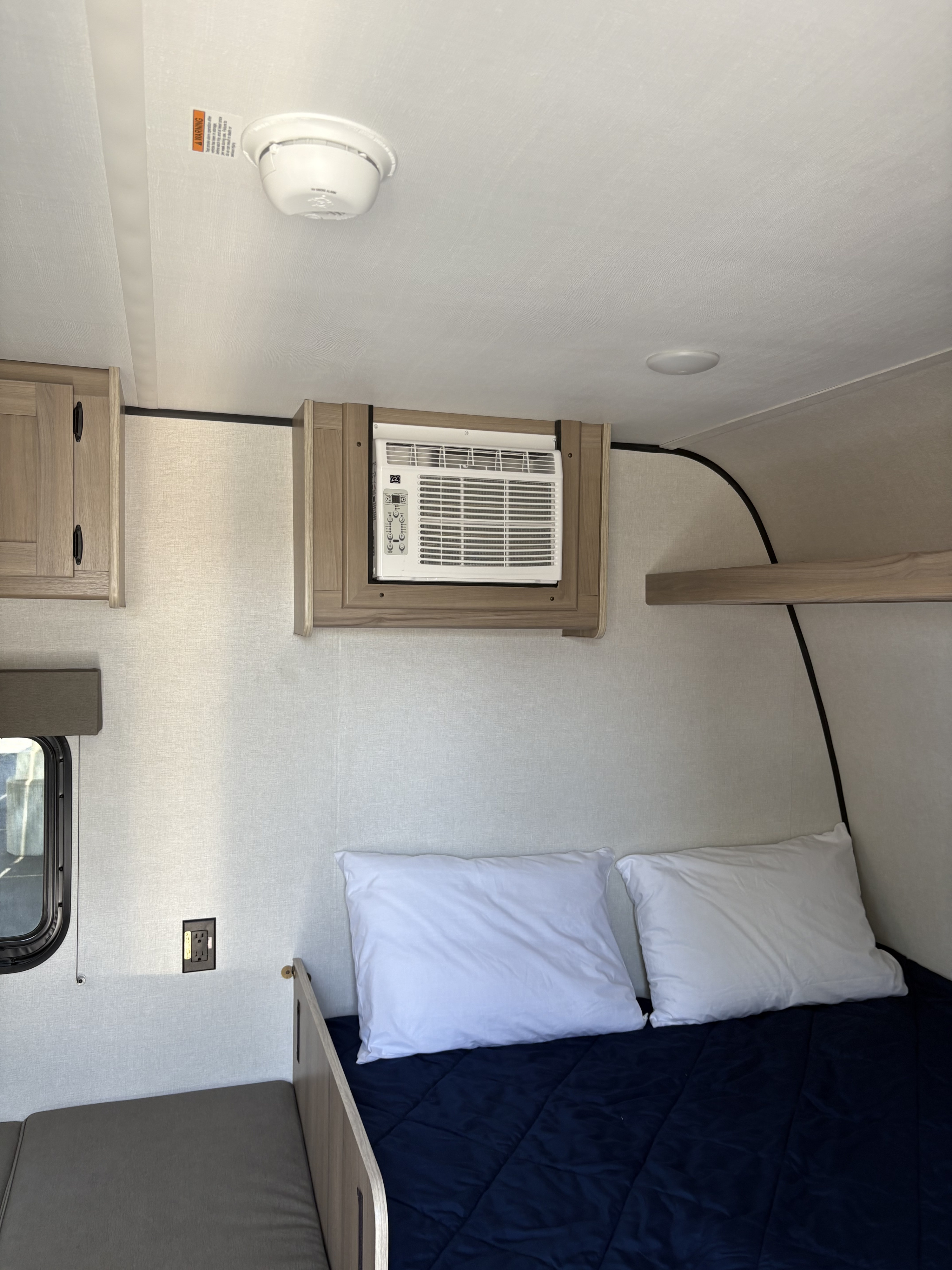Inside an RV rental: a cozy bed, window AC unit, and overhead storage. Smoke detector visible on ceiling.. Coleman 17B 2025