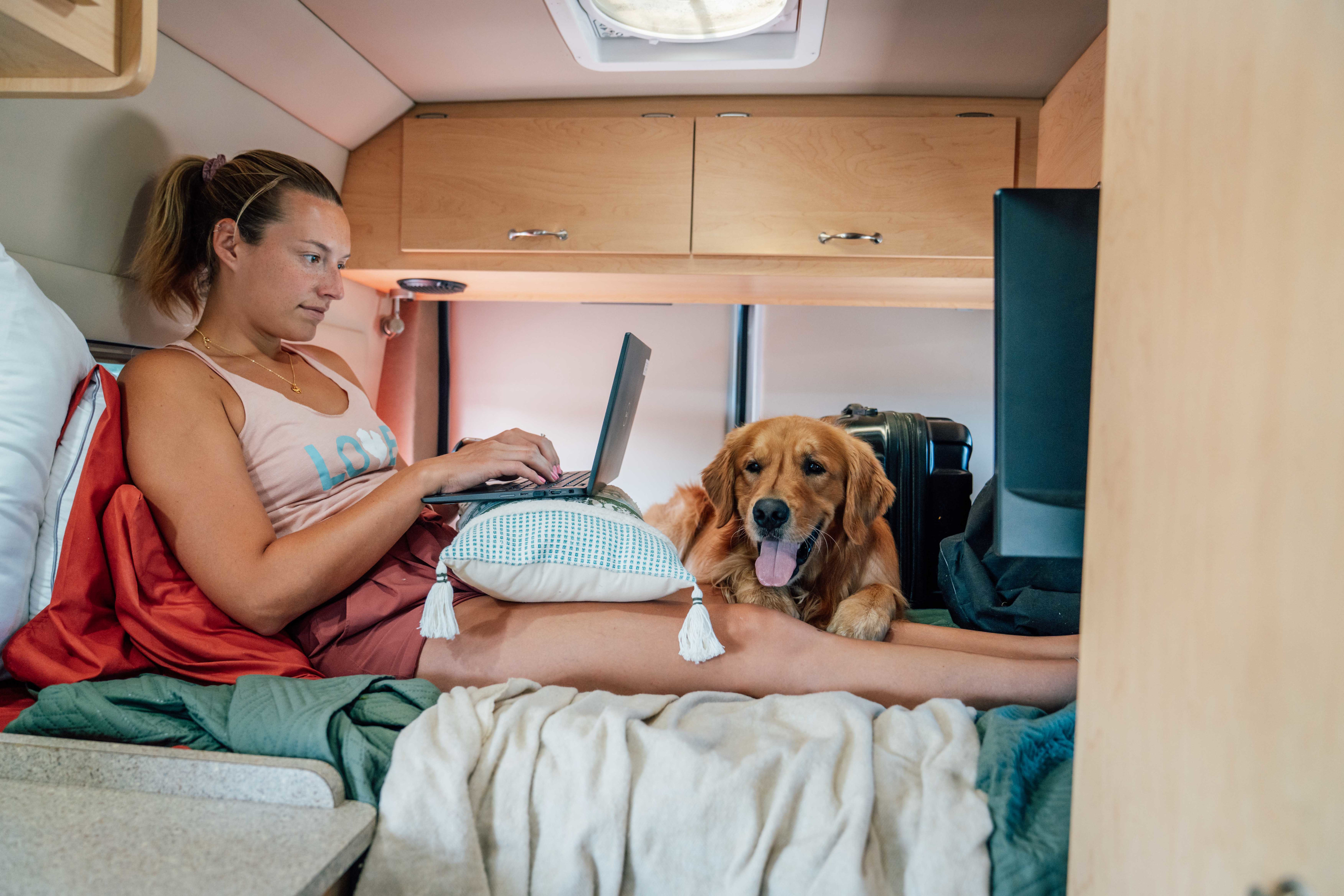 Cozy RV rental work setup: Woman on laptop with golden retriever by her side inside a camper van. Ideal for digital nomads.. Dodge Sprinter Van 2019