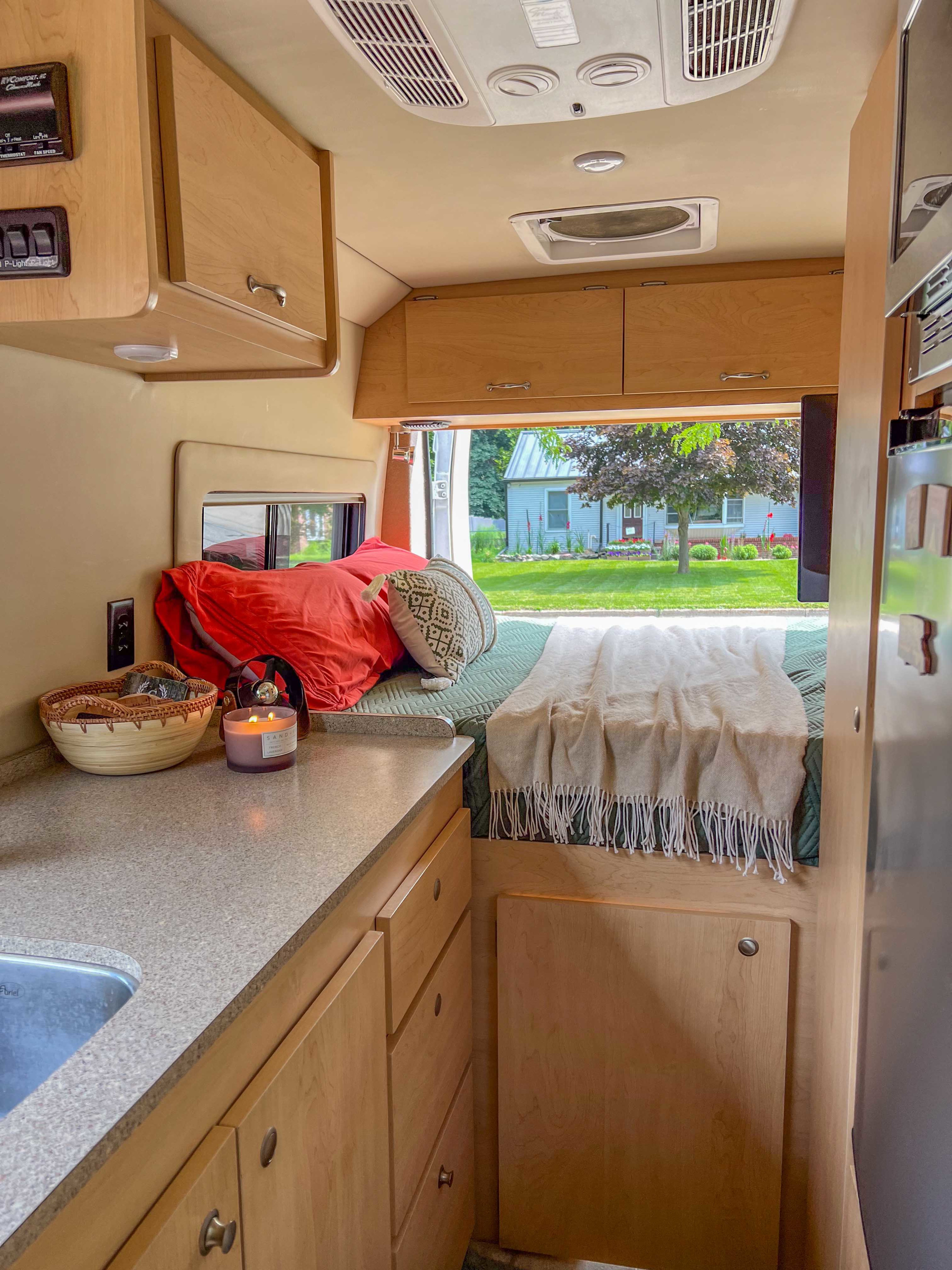 Cozy RV rental interior with a functional kitchen counter, comfortable bed, and a scenic backyard view from the large window.. Dodge Sprinter Van 2019