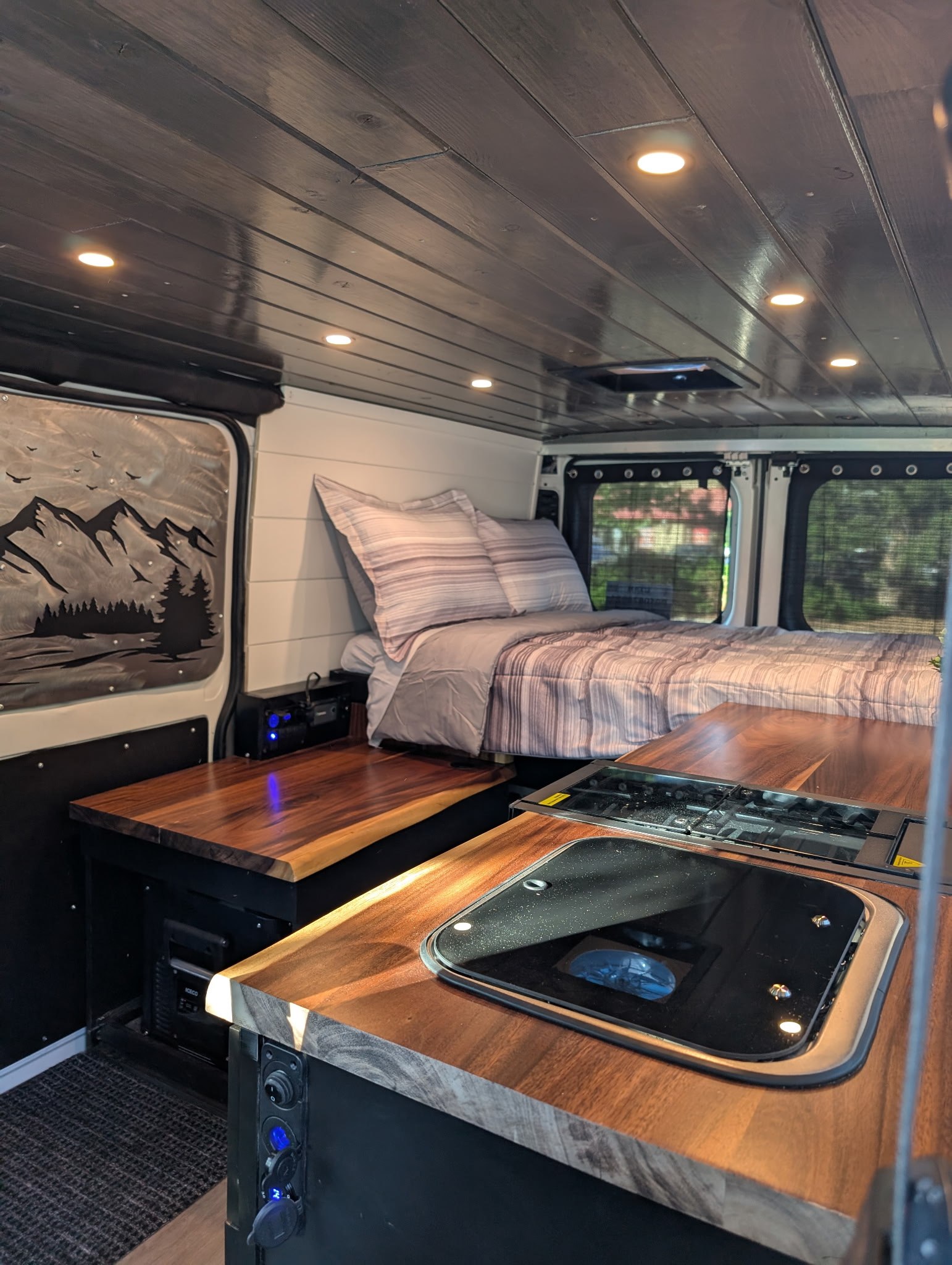 RV rental interior showing a comfortable bed, custom wood countertops, an integrated stove, and ceiling lights.. Custom Ram Promaster 1500 2017