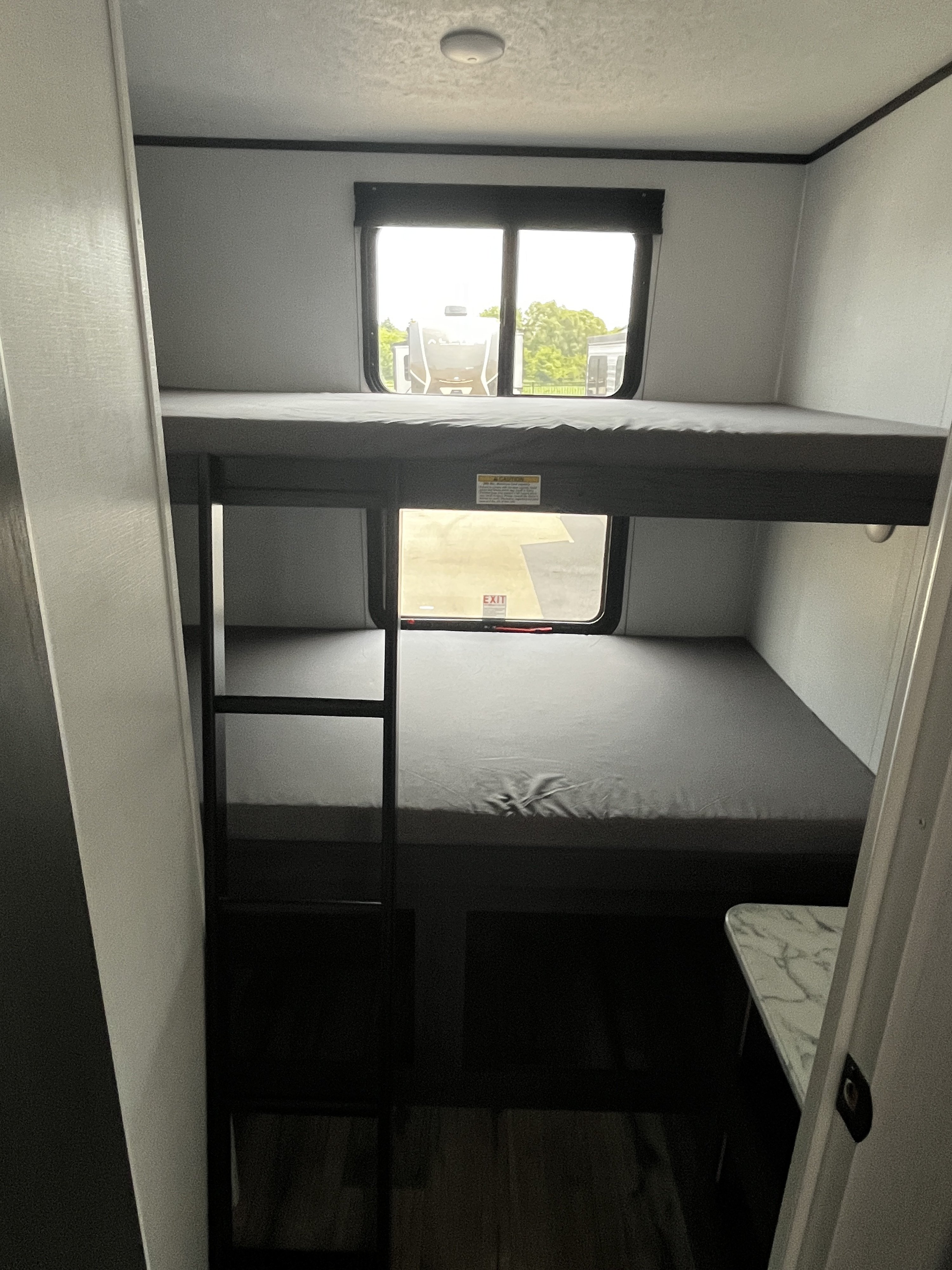 Inside an RV rental: a compact sleeping area with two gray bunk beds, a ladder, and large windows offering exterior views.. Dutchmen Aspen Trail 2025