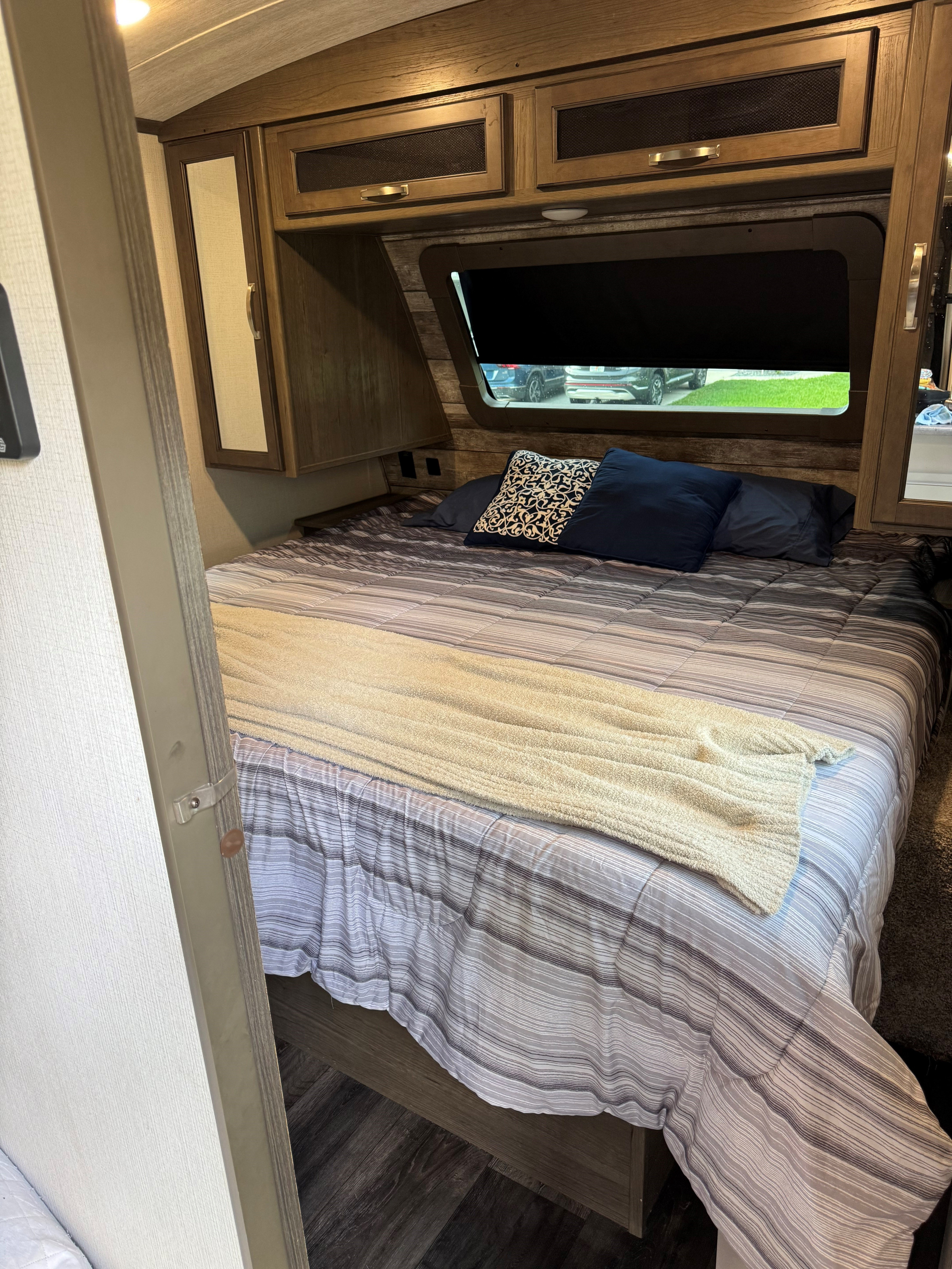 RV rental bedroom showing a striped bed, pillows, wood cabinets, and window with a view.. Keystone RV Cougar 2021