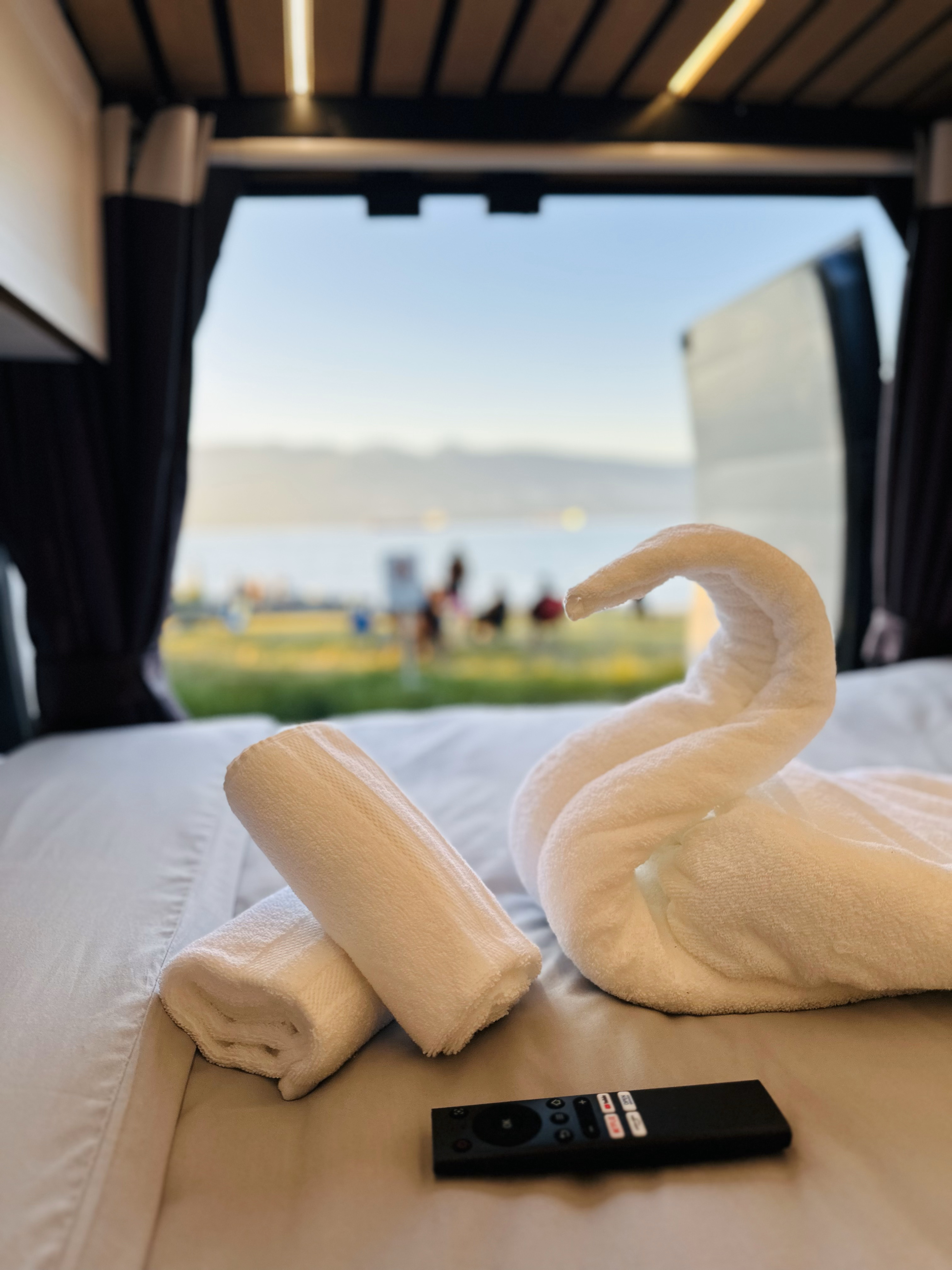 Enjoy the cozy RV rental bed with swan towel & remote, overlooking a serene lake view with people relaxing outdoors.. Dodge Promaster 3500 2016
