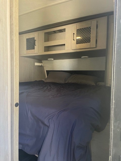 View into a cozy RV rental bedroom featuring a dark blue bed with pillows and light wood overhead cabinets.. Heartland RVs Mallard 2021
