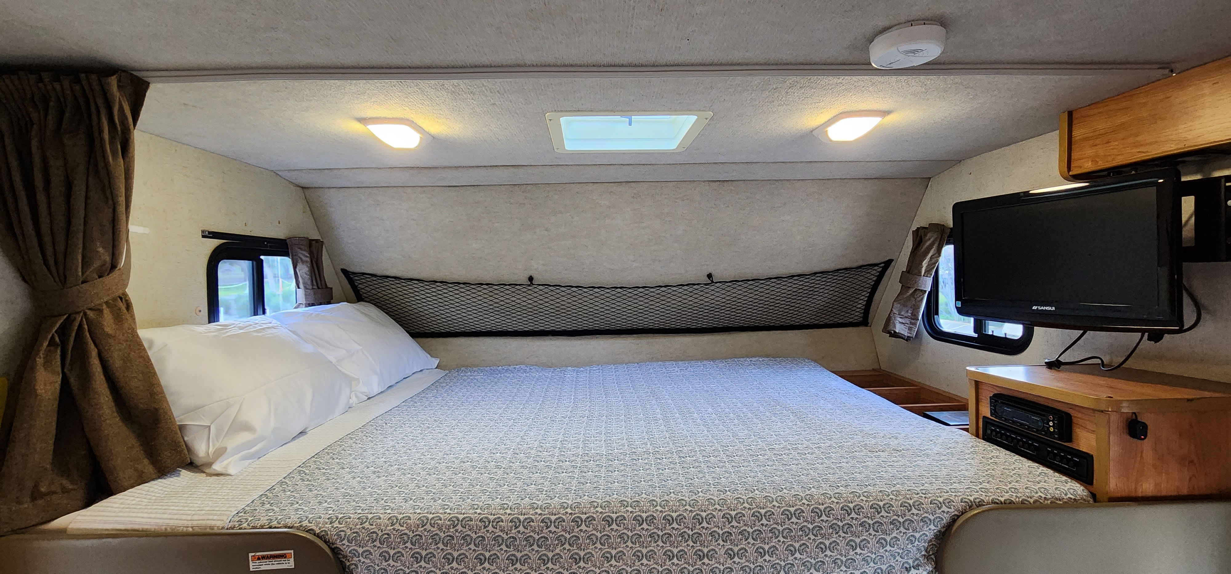 Inside an RV rental: A clean, inviting bed with white pillows, side windows, a mesh storage net above, mounted TV, and ceiling lights.. Jamboree Sport 2010