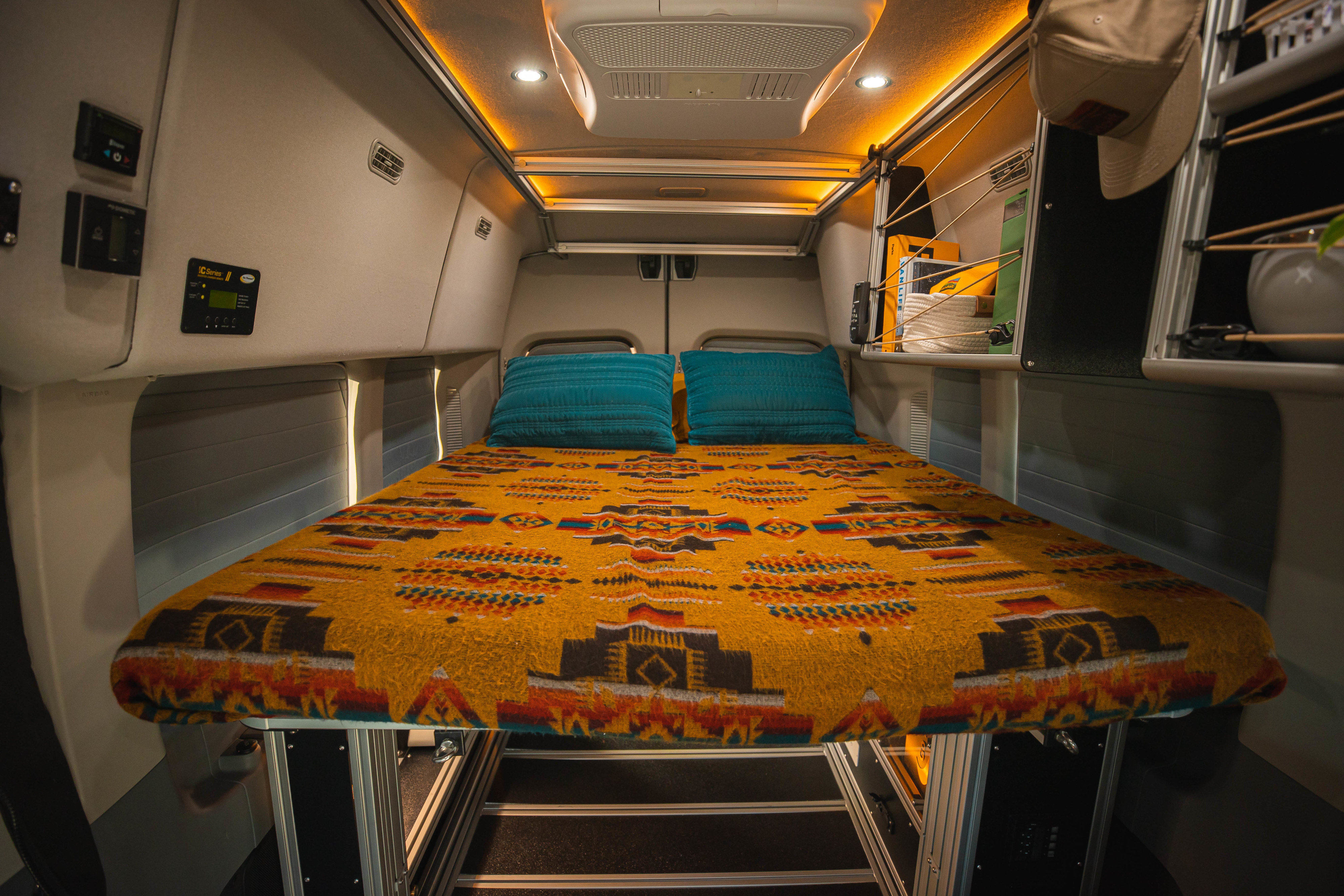 Cozy RV rental interior with a vibrant patterned bed, overhead lighting, and built-in storage, perfect for van life adventures.. Vandoit LIV 2019