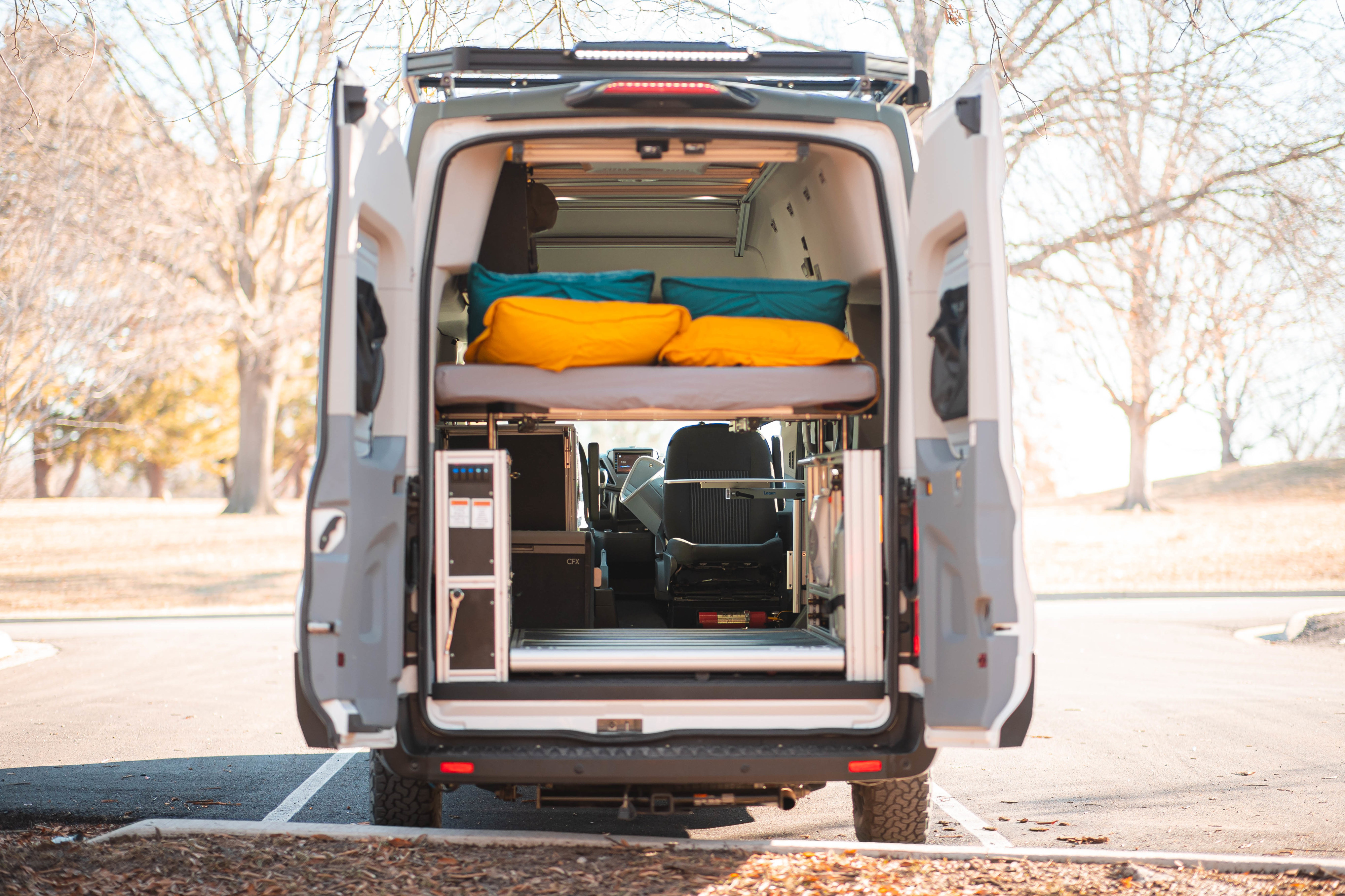 Open rear of an RV rental camper van reveals a raised bed, control panel, cooler, and passenger seat, perfect for road trips.. Vandoit LIV 2019