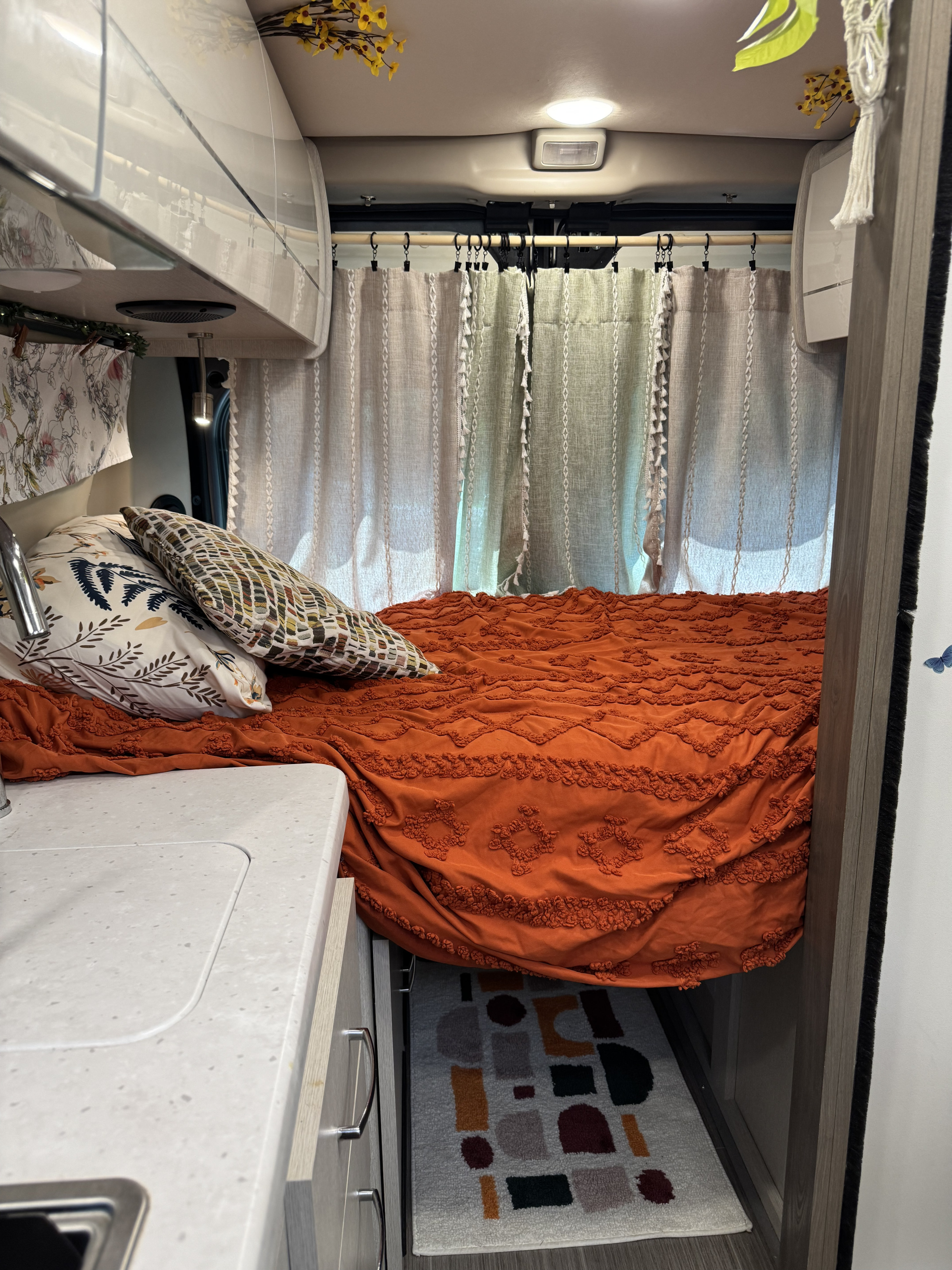 Cozy RV rental interior: orange bed with pillows, sunlit curtains. Counter & geometric rug visible, creating a homey feel.. Thor Motor Coach 18A Rize 2023