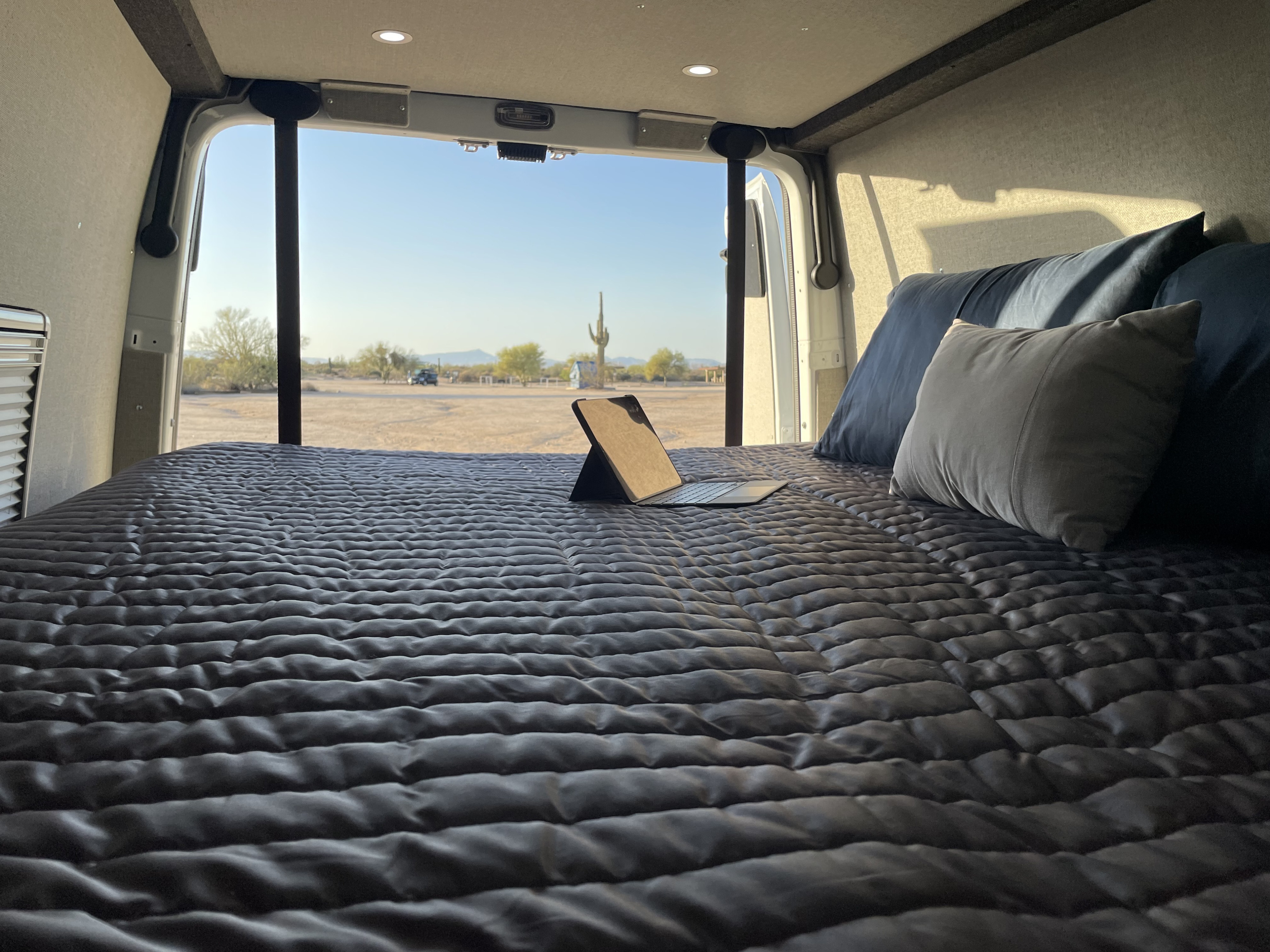 RV rental: Relax in a comfy bed in your camper van, enjoying desert views with saguaro cacti. Laptop on bed for convenience.. Mercedes Sprinter 2023