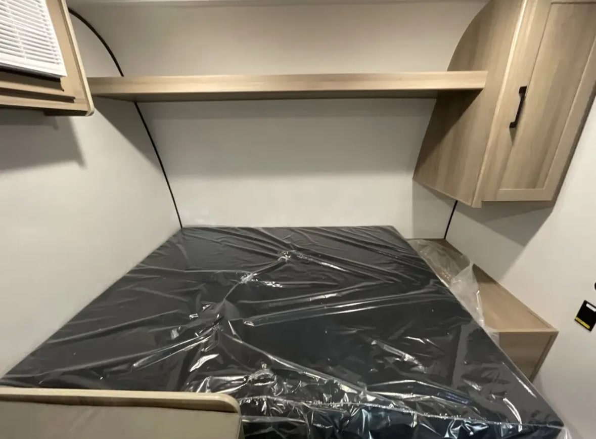 Compact RV rental sleeping area features a new, plastic-wrapped mattress, shelf, and cabinet overhead.. Other Other 2025