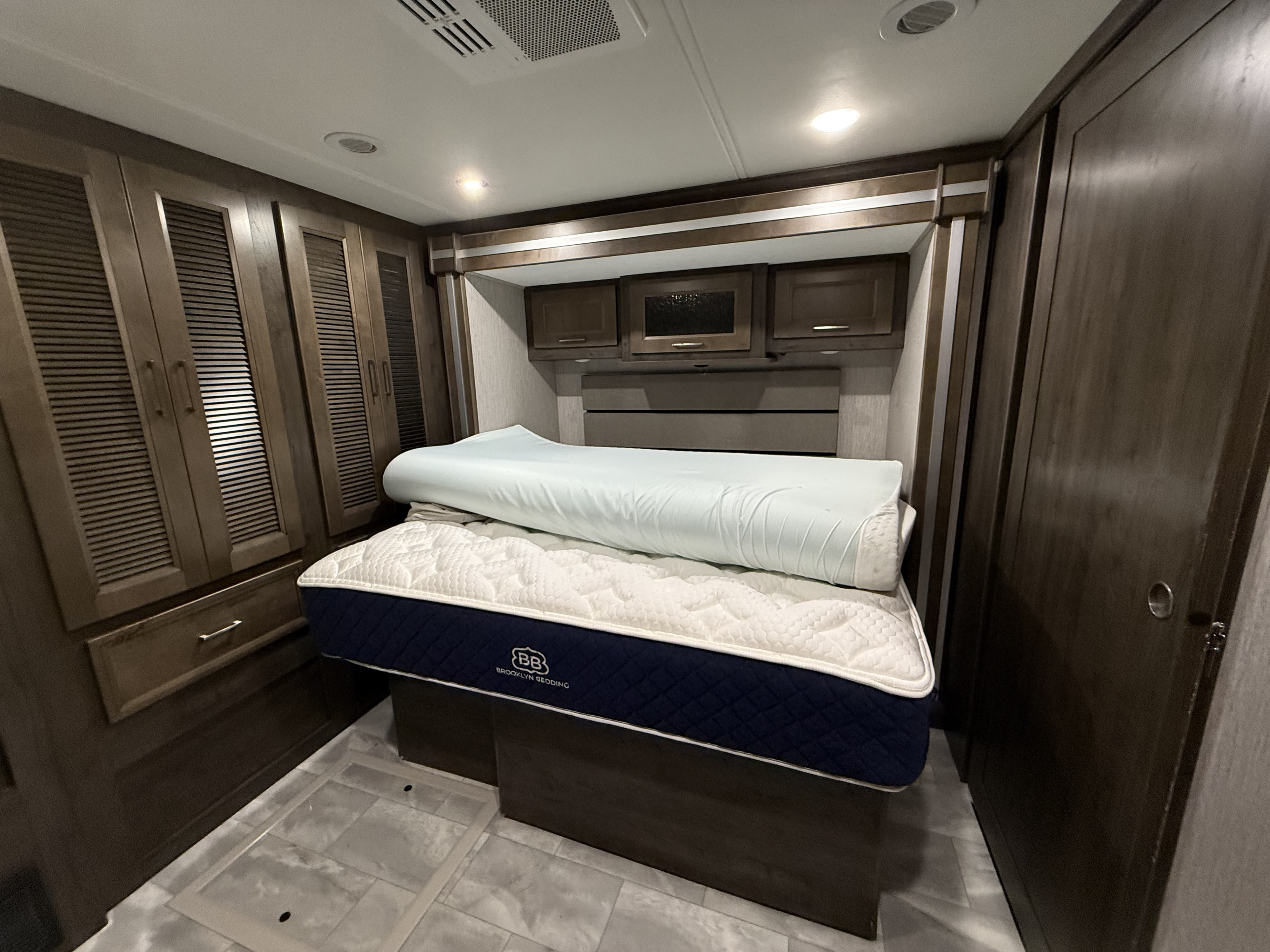 RV rental bedroom: a bed with a Brooklyn Bedding mattress, rolled topper, wood wardrobes, and overhead cabinets.. Coachman Sports coach 2021