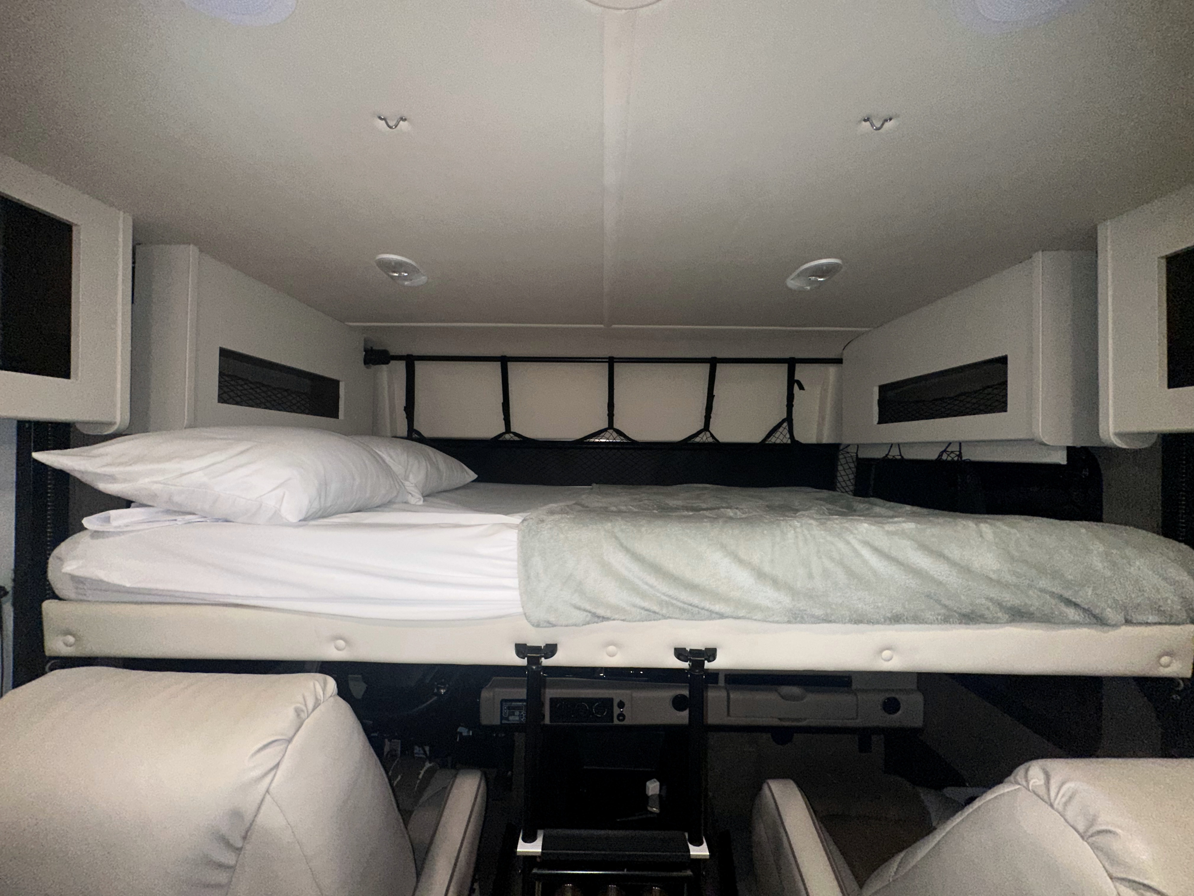 Cozy RV rental sleeping area: a neatly made bed with white linens, light grey blanket, overhead storage, and safety net.. Fleetwood Flair 2021
