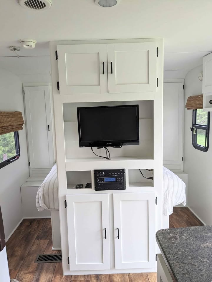 Modern RV rental interior with white built-in cabinetry, TV, and media center, leading to a cozy bed space.. Keystone RV Passport Grand Touring 2014