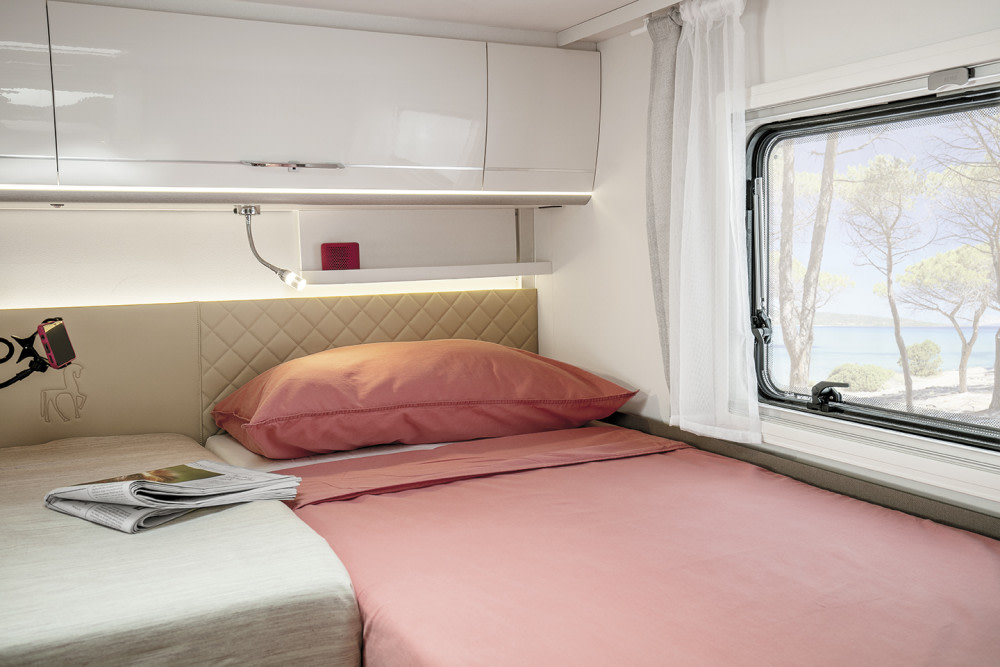 Cozy RV rental bedroom: dual beds, overhead storage, reading lamp, & a window with a beautiful beach view.. Etrusco I7400SBC 2025