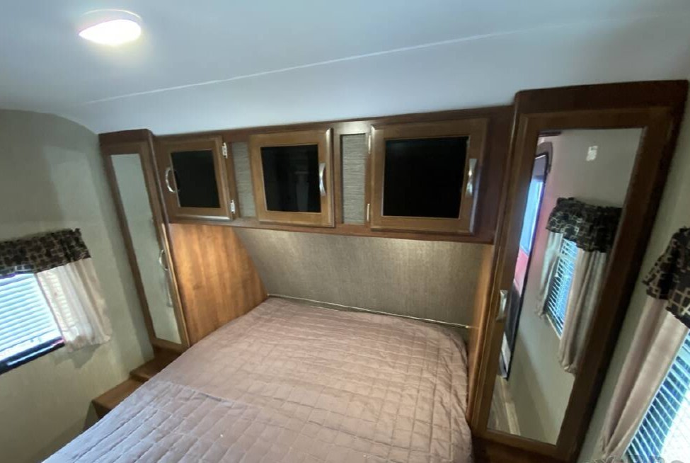 This RV rental bedroom features a comfy bed, overhead storage cabinets, and a full-length mirror, perfect for travel trailer adventures.. Keystone RV Passport Grand Touring 2018