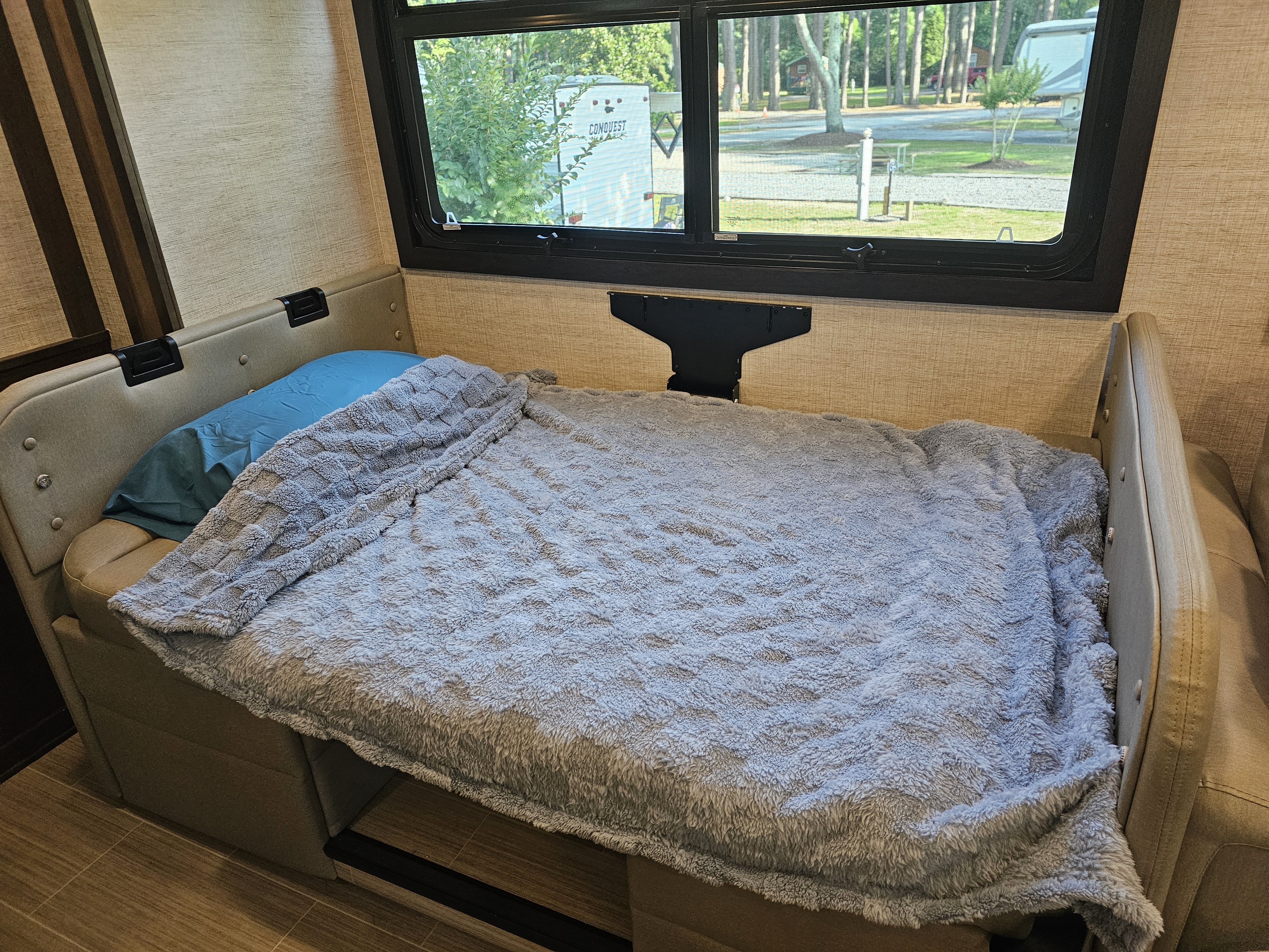 Inside an RV rental: a comfy dinette bed with blue pillow and grey blanket, overlooking a serene campground through the window.. Entegra Coach Vision XL 2022