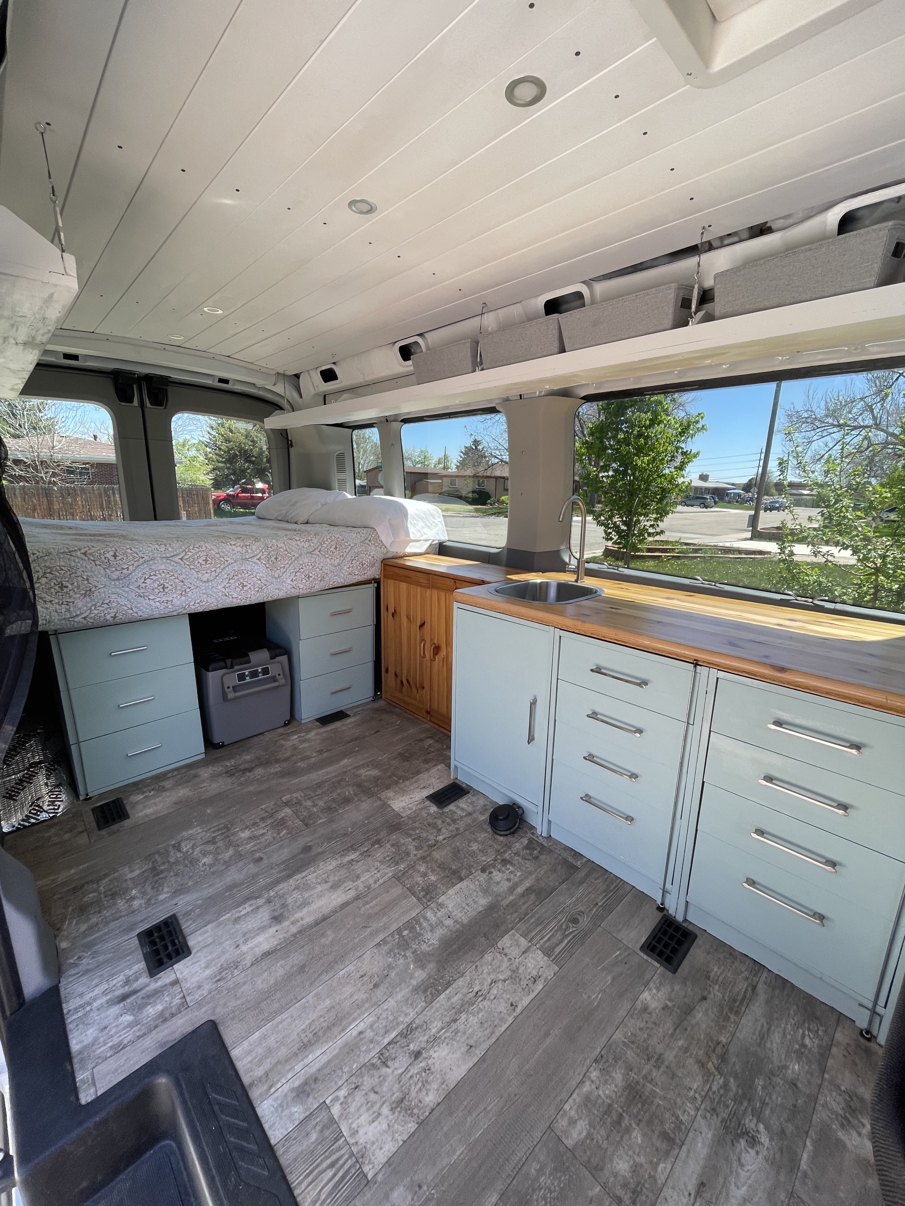 Bright RV rental interior: light blue kitchen, wood-look floor, cozy bed, and overhead storage. Ideal for travel.. Ford Transit Custom 2017