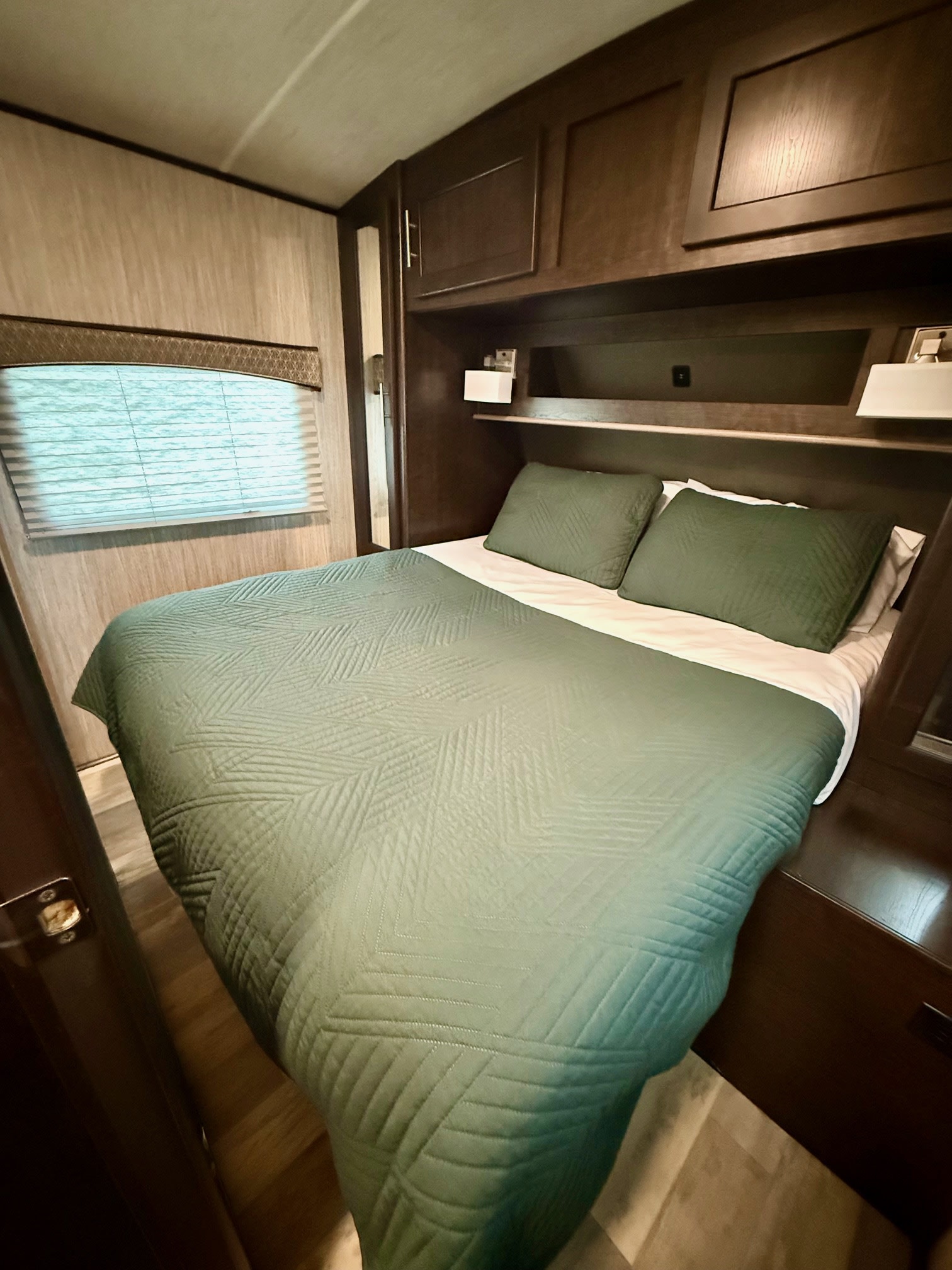 View of a compact RV rental bedroom. A green-quilted bed sits below dark wood cabinetry and a window with blinds.. Cruiser RV Fun Finder Xtreme Lite 2019