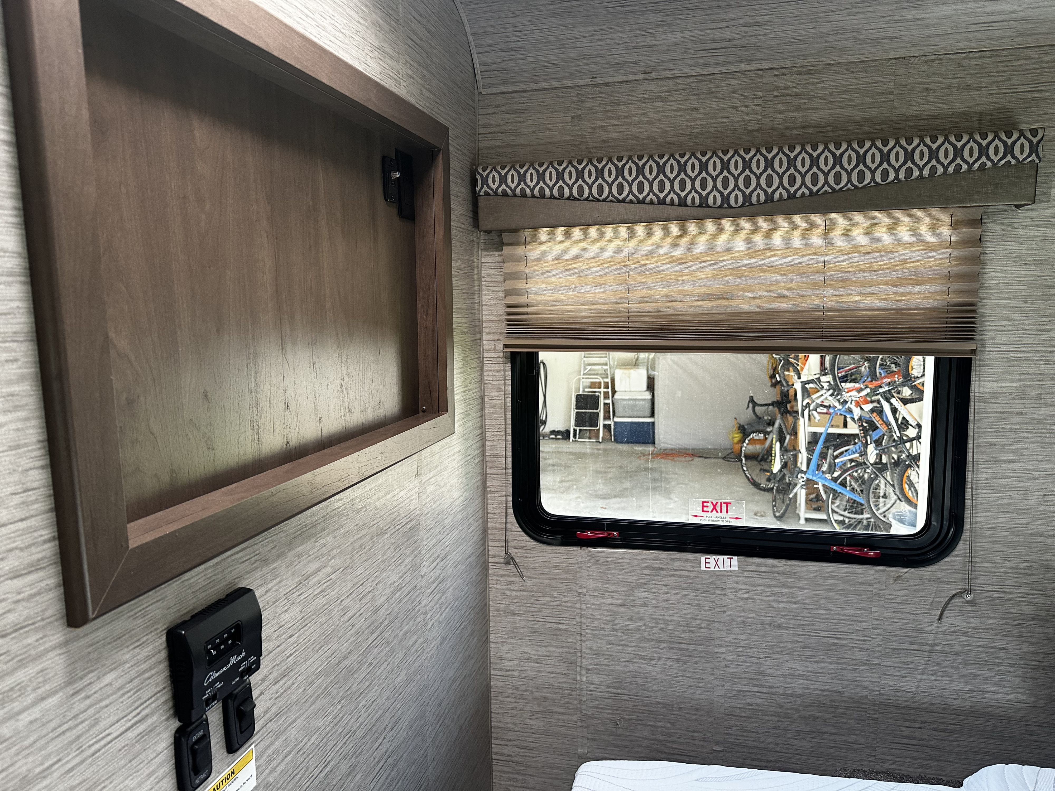 RV rental interior: A window reveals bikes in a garage, alongside a control panel and wooden cabinet. Emergency exit marked.. Thor Freedom Elite 2021